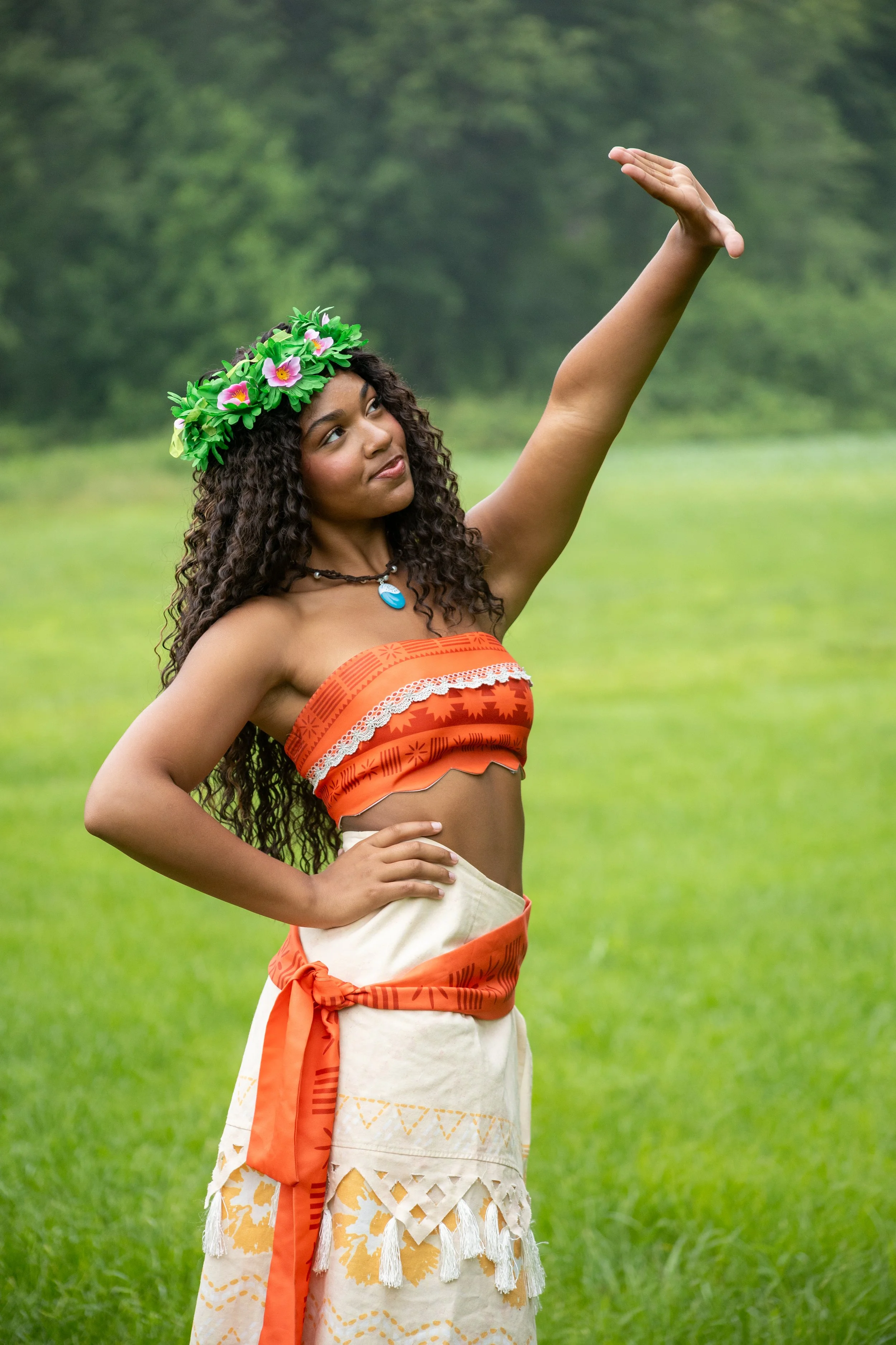 Woman in traditional Hawaiian attire standing outdoors in a grassy field, wearing a flower crown, and posing with her right arm raised.