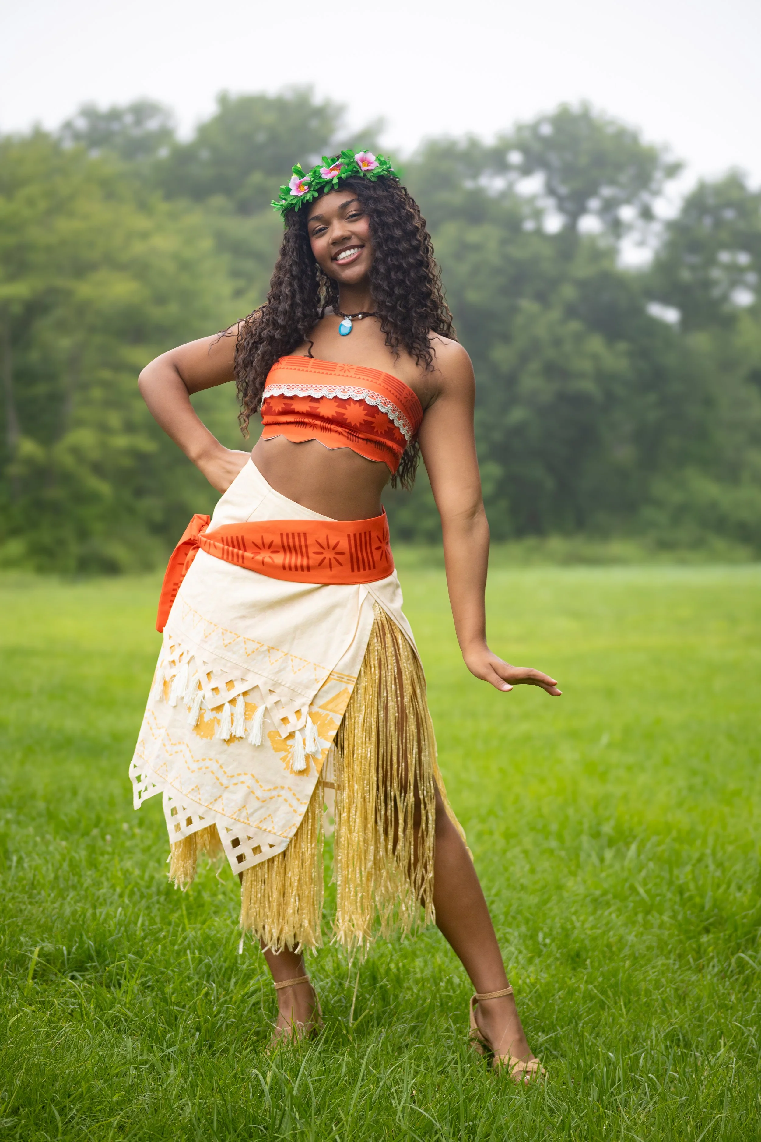 A woman dressed in traditional Hawaiian attire, wearing a orange and white skirt, an orange crop top, a flower crown, and dancing outdoors on green grass with trees in the background.