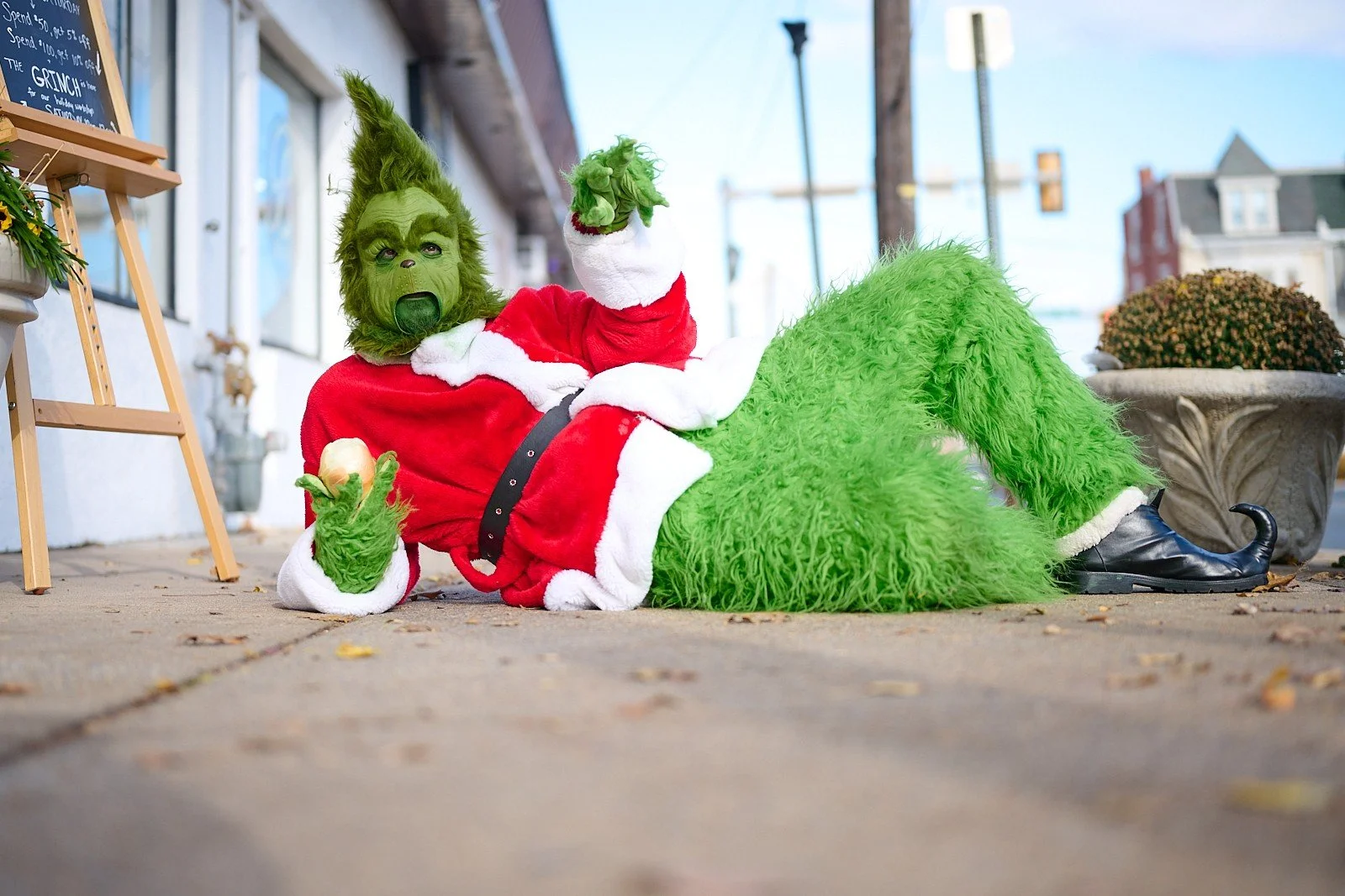 A person dressed as the Grinch, with green furry face and costume, lying on the sidewalk in a Christmas Santa outfit, holding an onion, with one hand raised. The background shows buildings, a streetlight, and a potted plant.