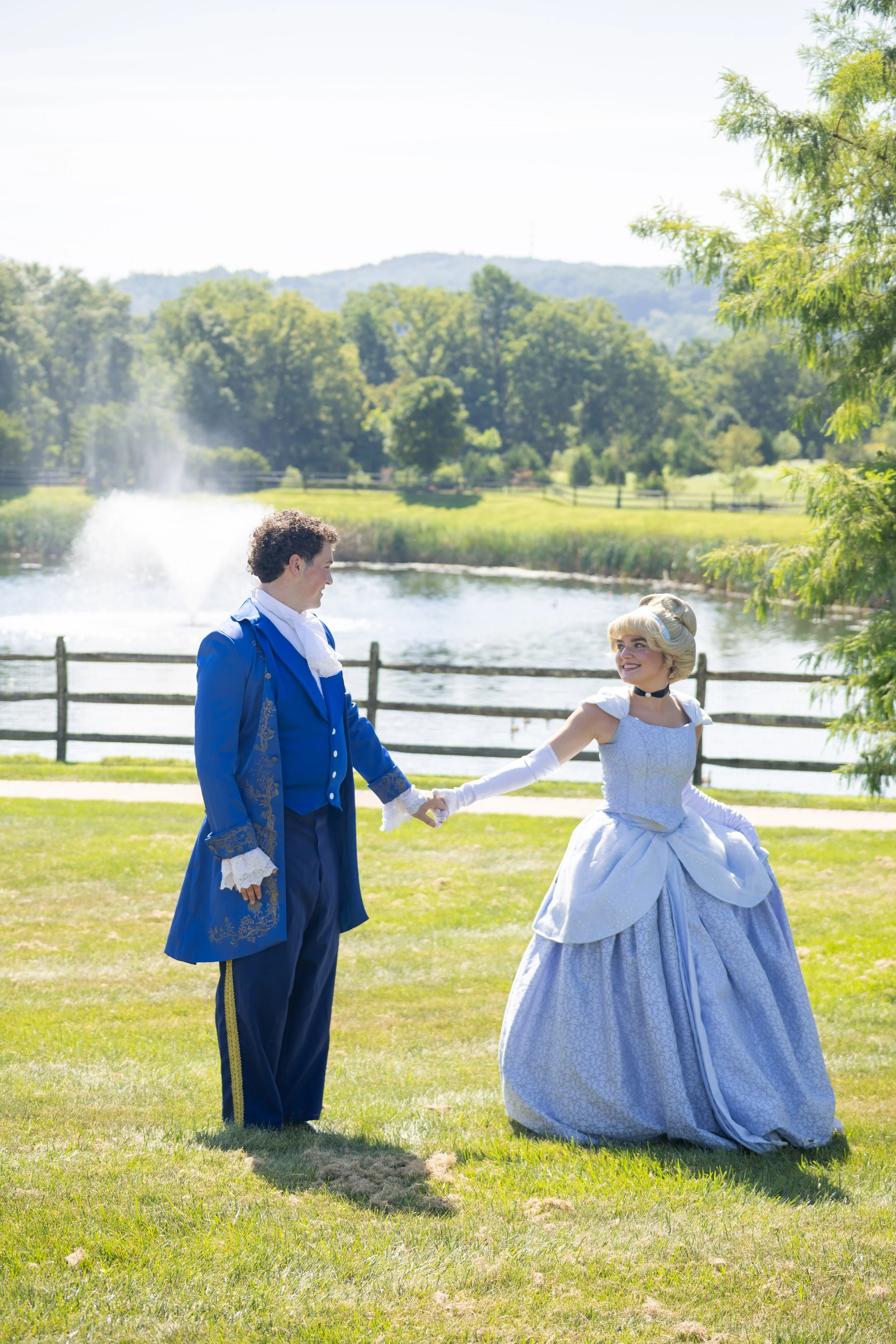 Couple dressed in historical costumes holding hands in a park by a lake with fountain, trees, and hills in the background on a sunny day.
