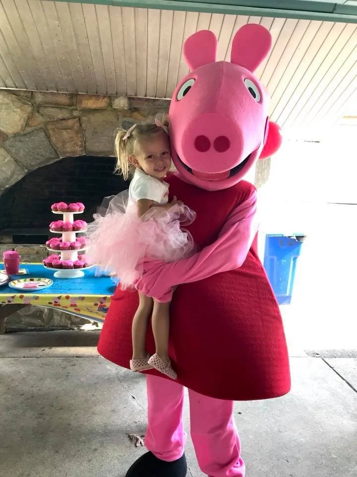 A young girl in a pink tutu and white shirt being embraced by a person in a Peppa Pig costume at a children's party with a table of pink treats and decorations in the background.