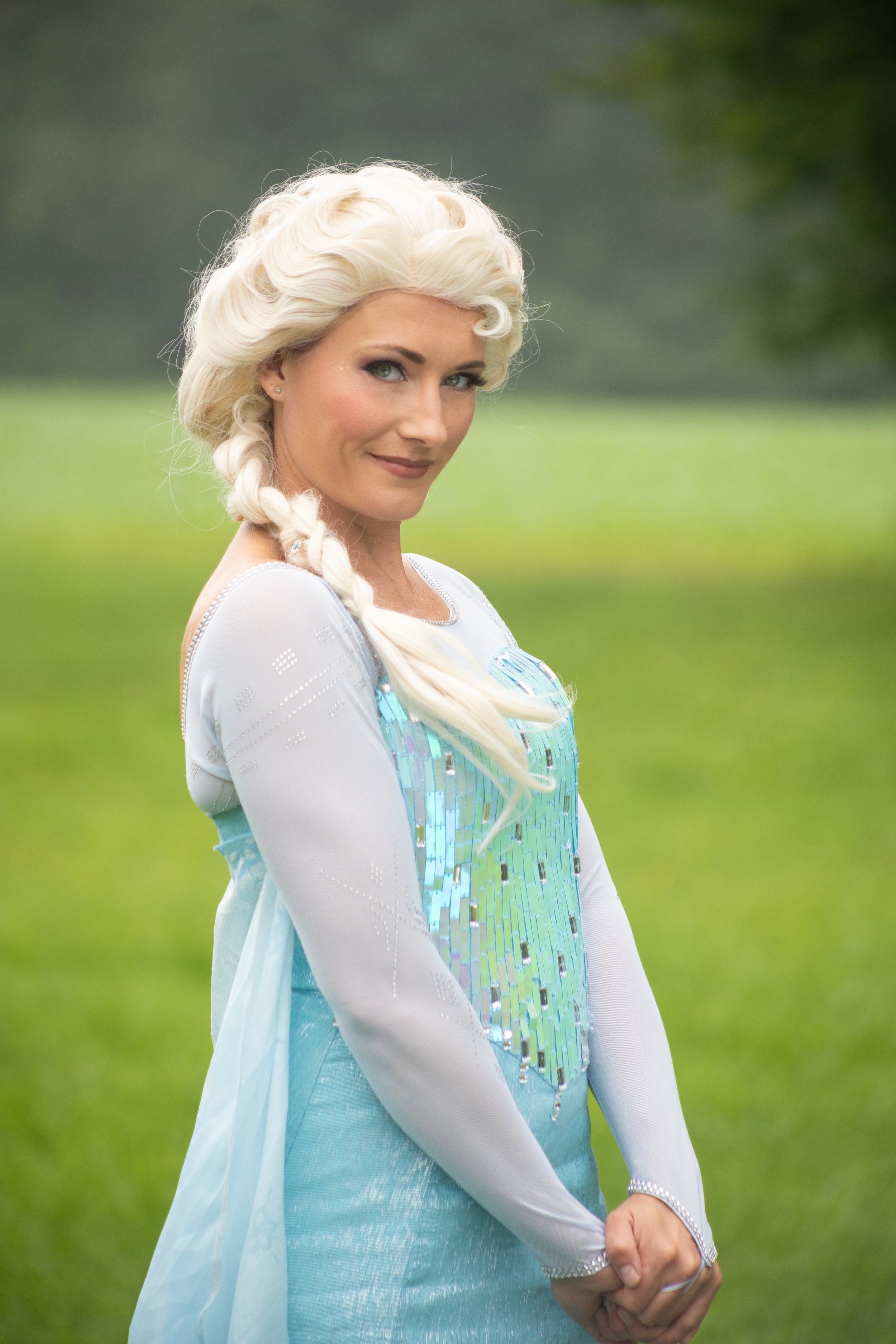 A woman dressed as a fairy tale princess with platinum blonde hair in a braid, wearing a light blue dress with shiny embellishments, standing outdoors on a grassy field, inspired by Queen Elsa.