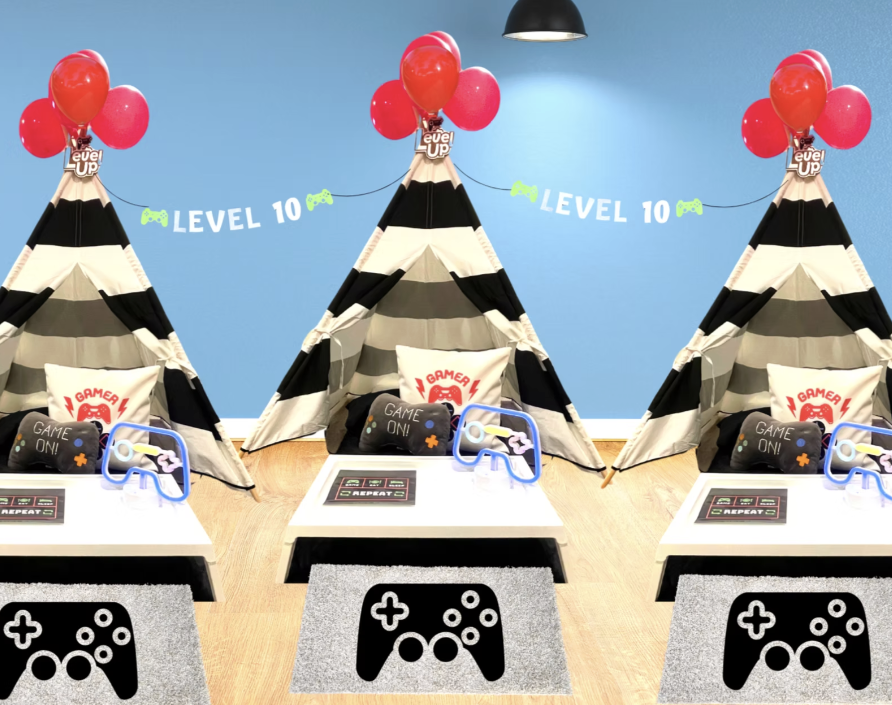 A gaming setup with three black and white striped tent-like structures decorated with red balloons and gaming-themed banners that say 'LEVEL 10' and 'LEVEL UP.' In front, there are gaming accessories, including pillows with game controllers, small ta