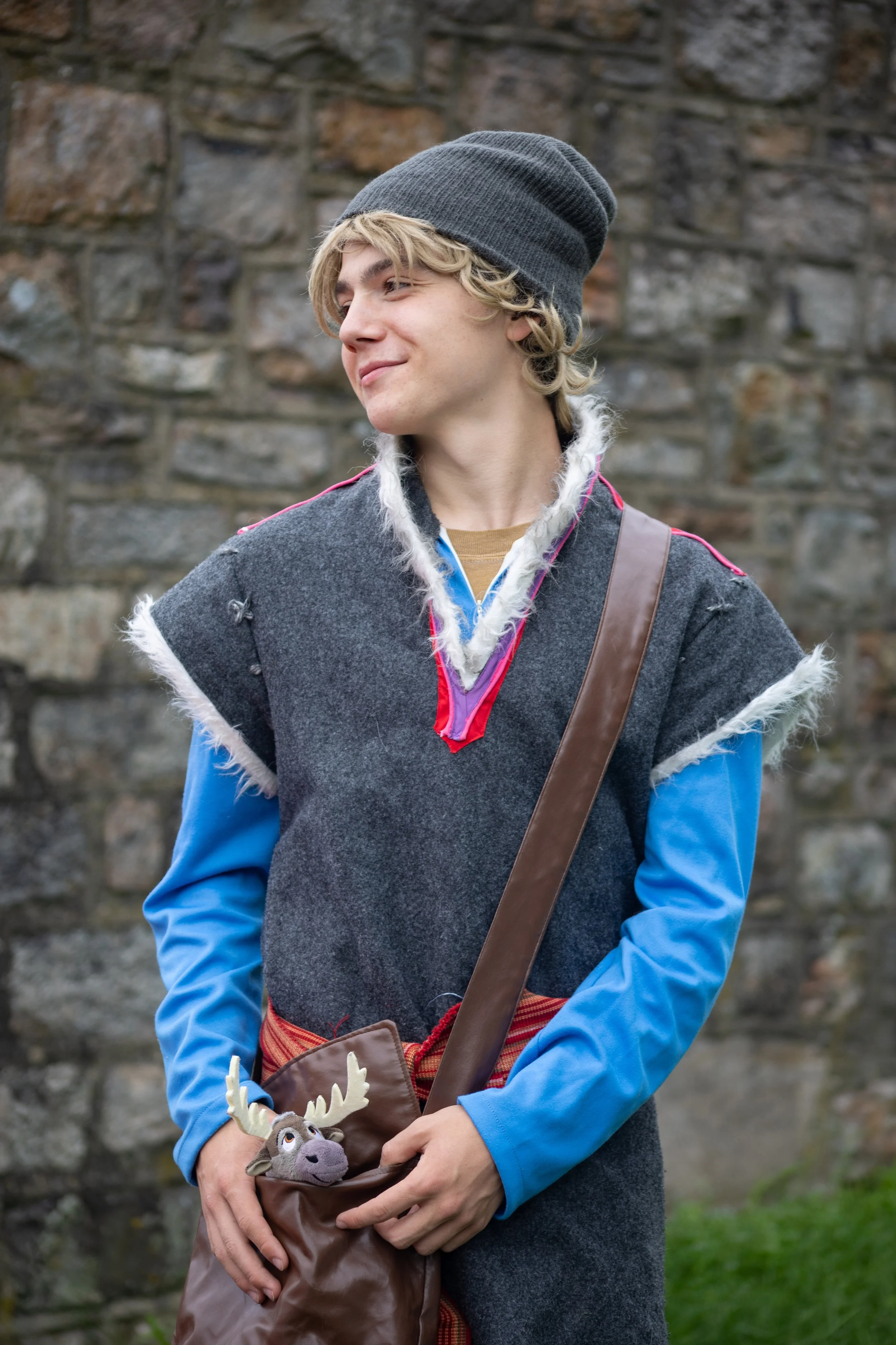 A young man in costume standing outdoors in front of a stone wall, wearing a gray beanie, a fleece-lined vest, a blue shirt, and carrying a brown bag with a plush reindeer, inspired by Kristoff and Sven from Frozen.