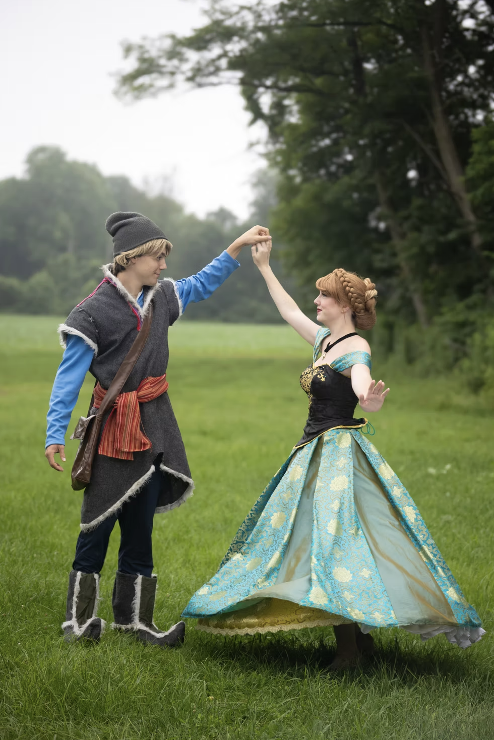 A young man and woman dressed in medieval or fantasy costumes dancing outdoors in a grassy field with trees in the background.