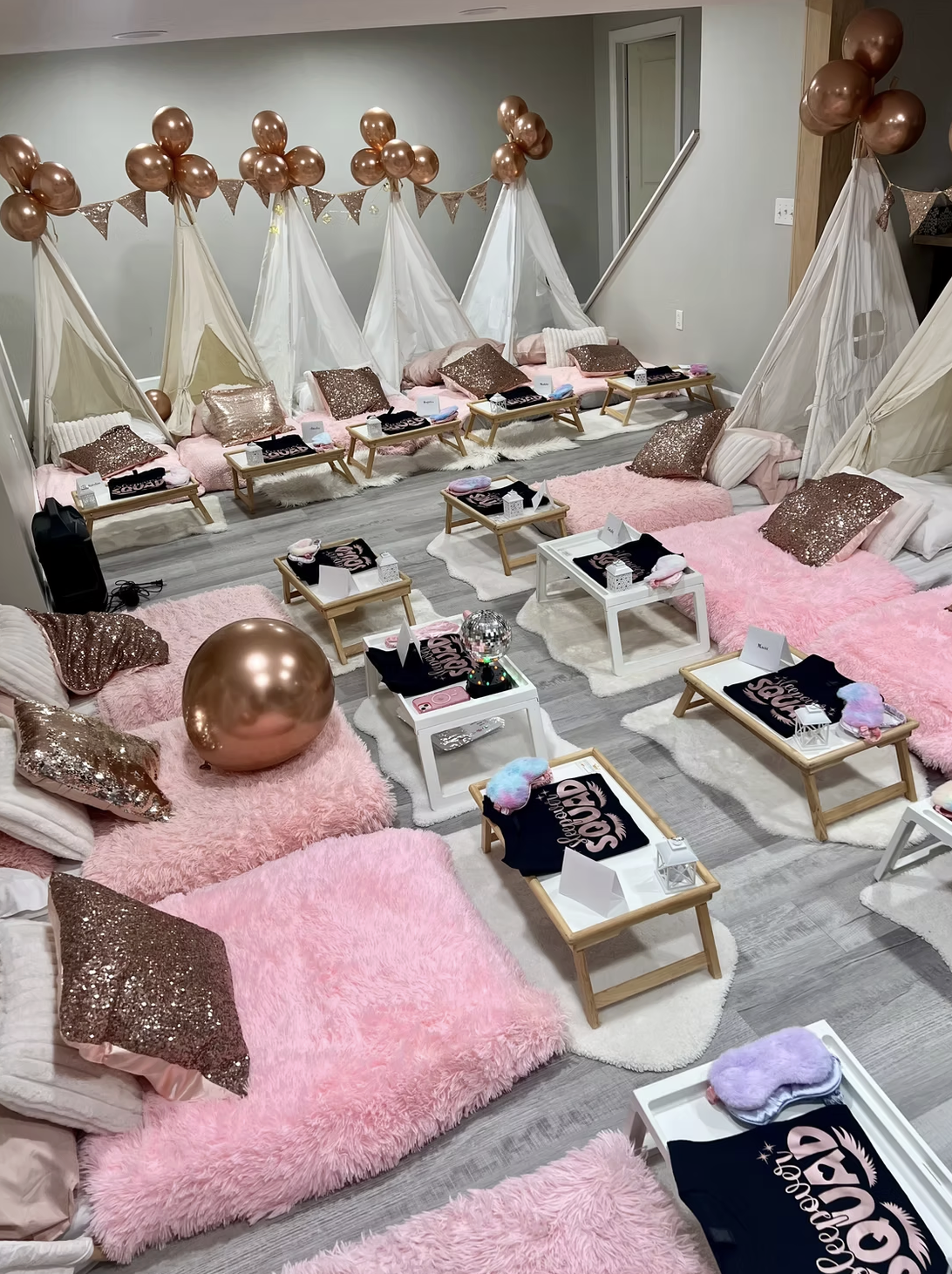 A cozy indoor space decorated for a sleepover or girls' night. There are multiple beds with pink fluffy blankets, glittery rose gold pillows, and small wooden beds with personalized t-shirts. The area features pink shaggy rugs, white tables with decorative items, balloons, and teepees with bunting in the background.
