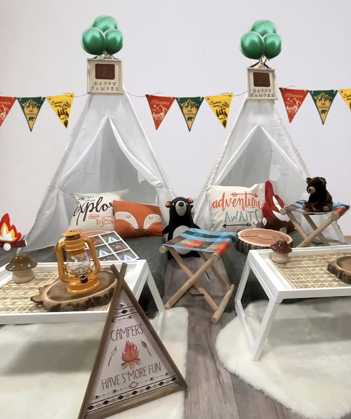 Campground themed indoor camping setup with miniature tents, plush animal pillows, camping decor, and signs that say "Campers have s'more fun" and "Explore, Adventure, Awaits".