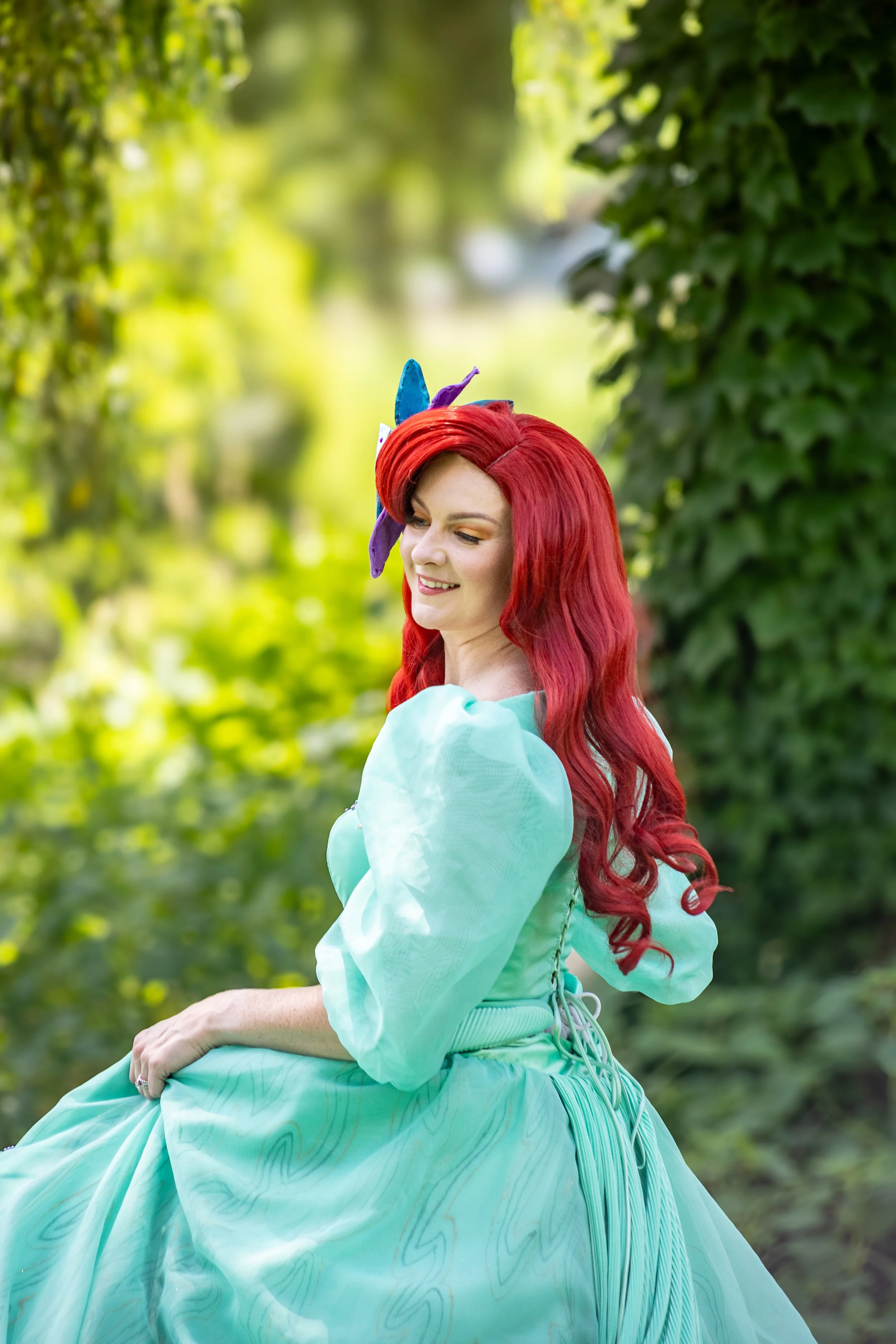 A woman with long, wavy red hair wearing a light blue dress with puffed sleeves, sitting outdoors amidst greenery, smiling.