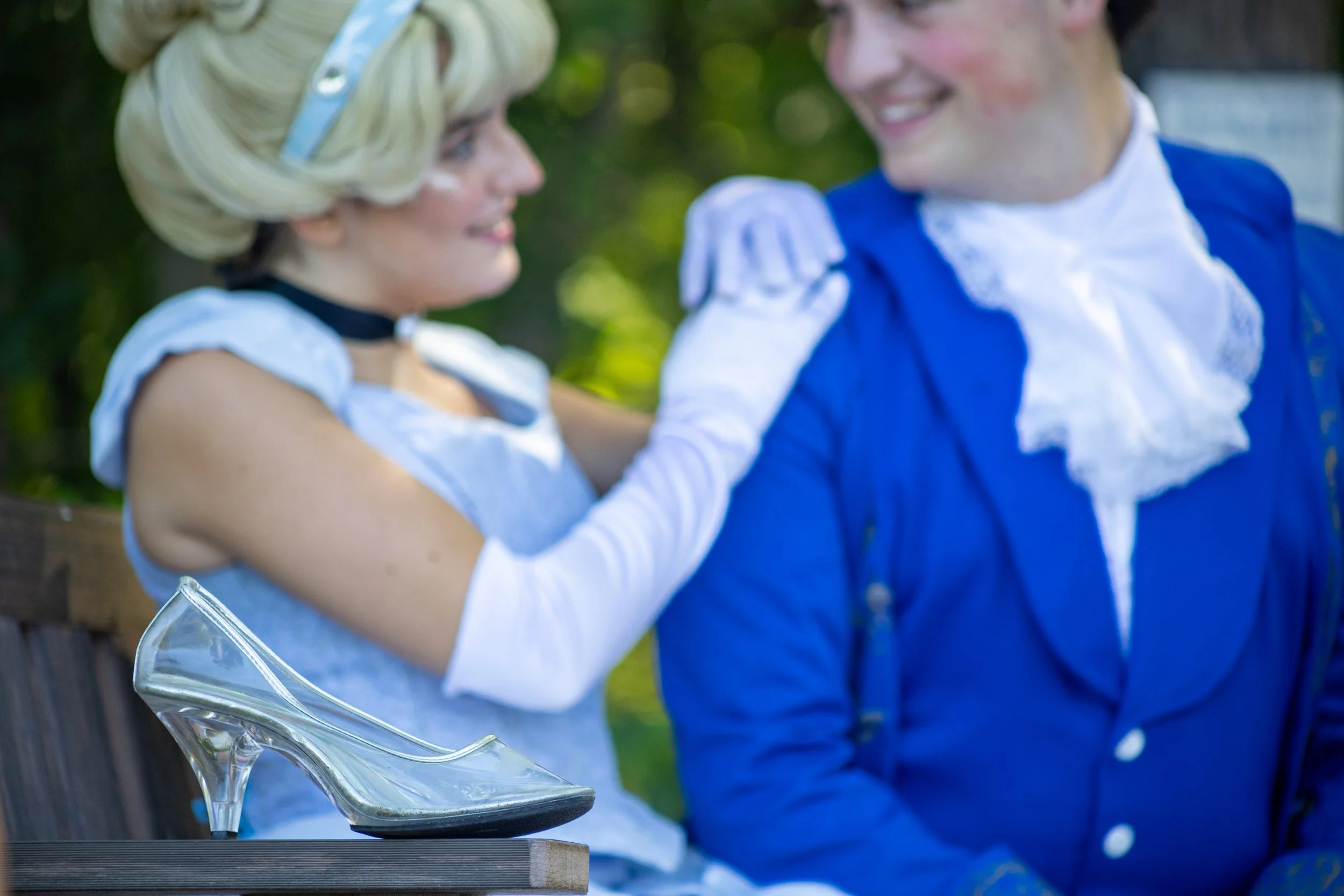 A woman and a man dressed in costumes inspired by historical European fashion, smiling at each other while sitting outdoors. A clear high-heeled shoe is placed on a bench in the foreground.