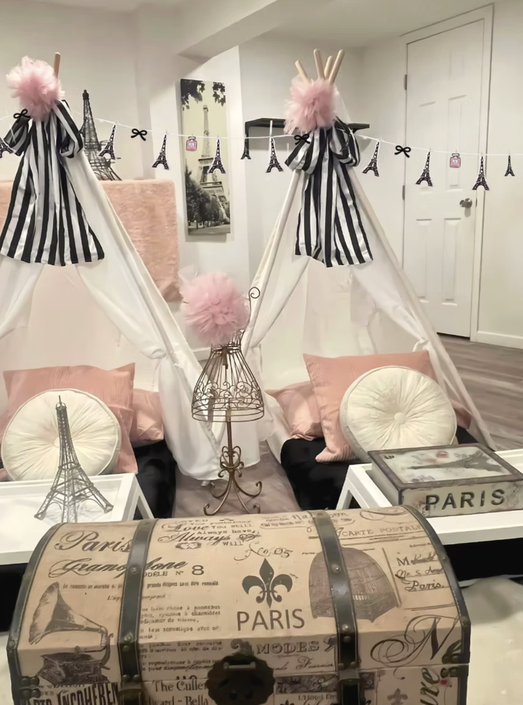 Decorative Paris-themed decor with a miniature teepee tent, Eiffel Tower images, pink fluffy pom-poms, striped banners, pink pillows, a vintage-style chest, and framed Paris photos in a cozy indoor setting.