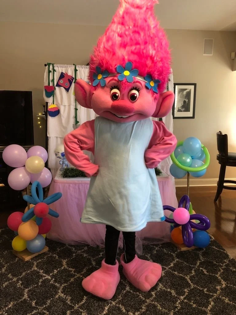 A colorful troll character with pink hair, big eyes, and pink feet, standing in a decorated party room with balloons and a pink and white themed backdrop.