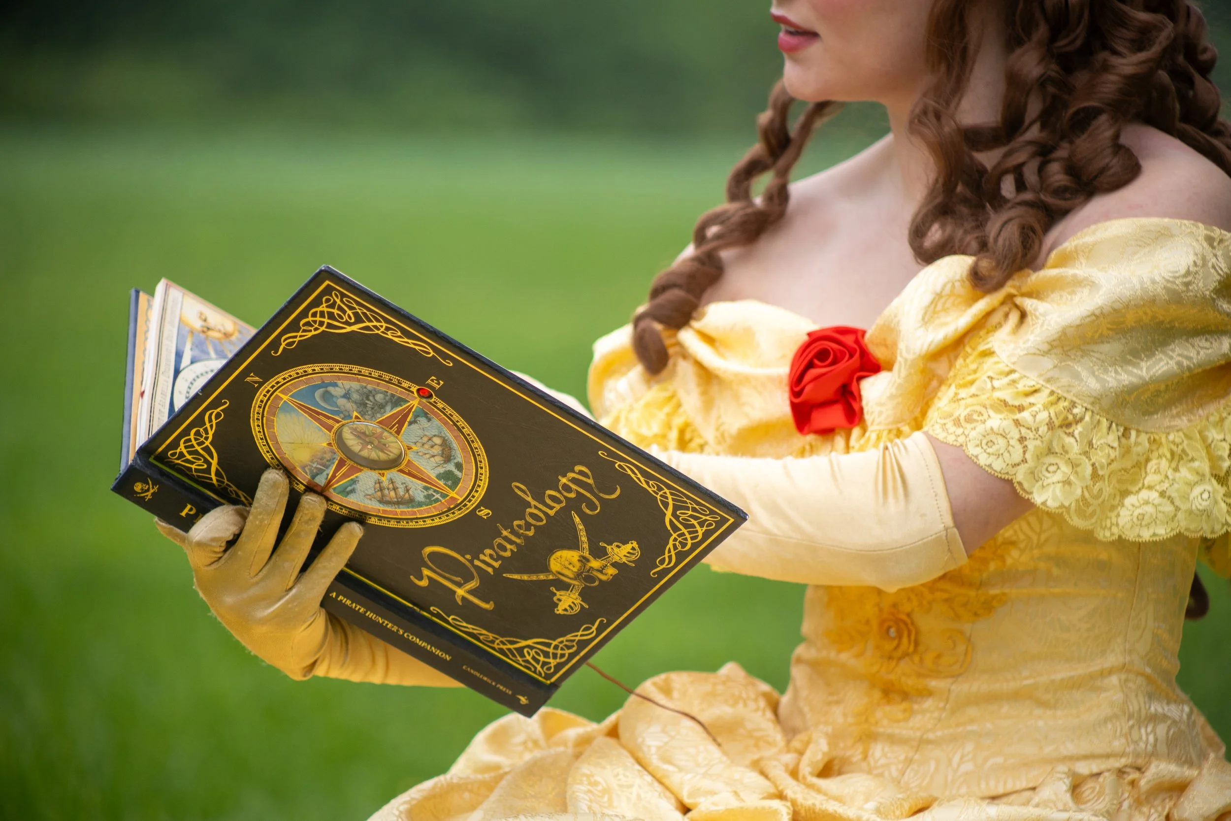 A woman dressed as Belle from Beauty and the Beast is holding an illustrated pirate dictionary with gold lettering and a compass on the cover, outdoors on a grassy background.