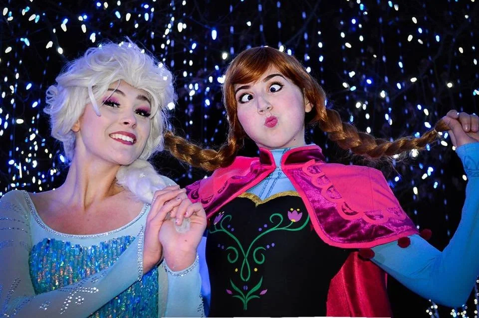 Two women dressed as Disney princesses Elsa and Anna from Frozen, posing against a backdrop of blue and white fairy lights.