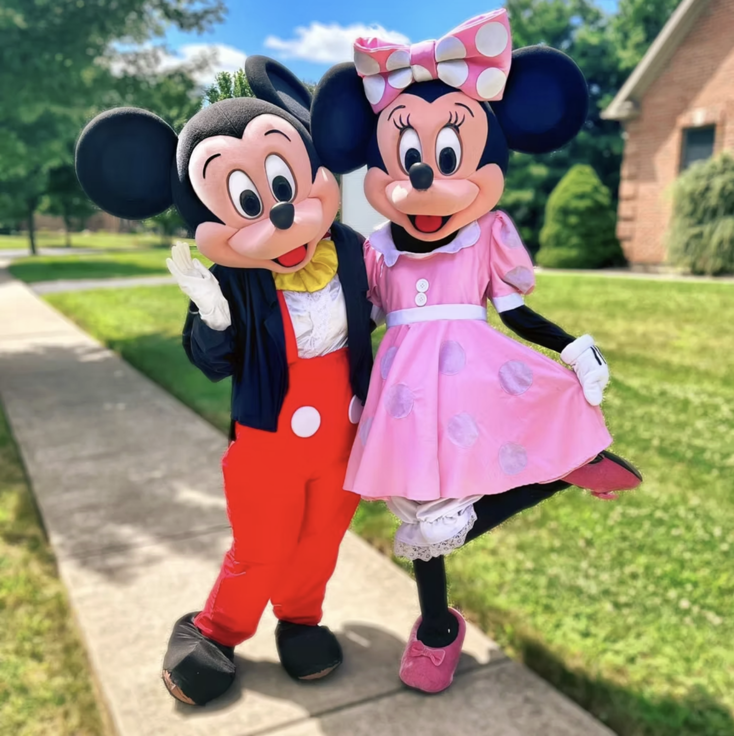 Costumed characters of Mickey Mouse and Minnie Mouse outdoors on a sidewalk with houses and trees in the background.
