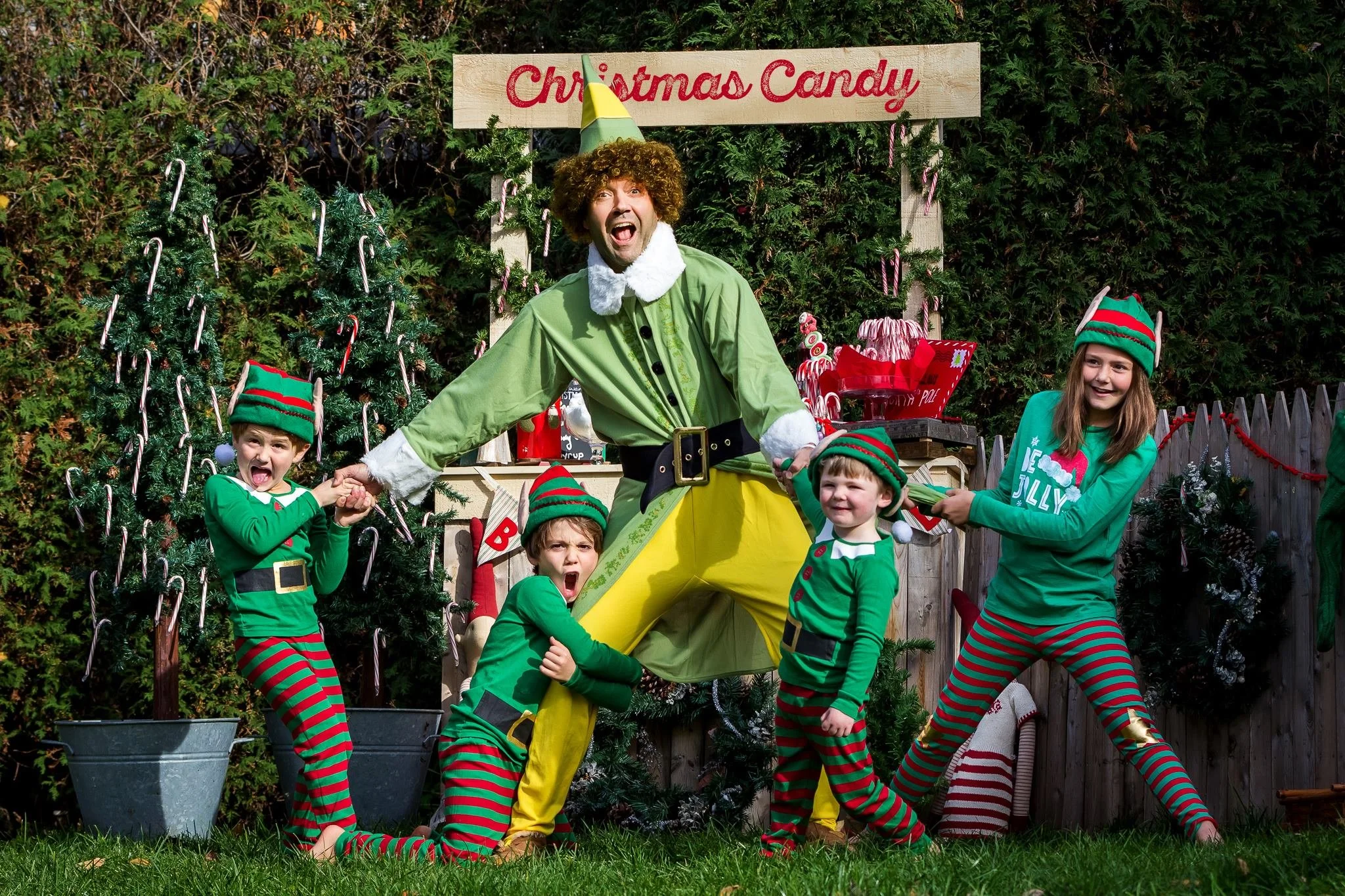 A person dressed as Buddy the elf in a green shirt and yellow pants holding hands with four children wearing elf costumes, standing in front of a Christmas display with Christmas trees and a sign reading 'Christmas Candy'.