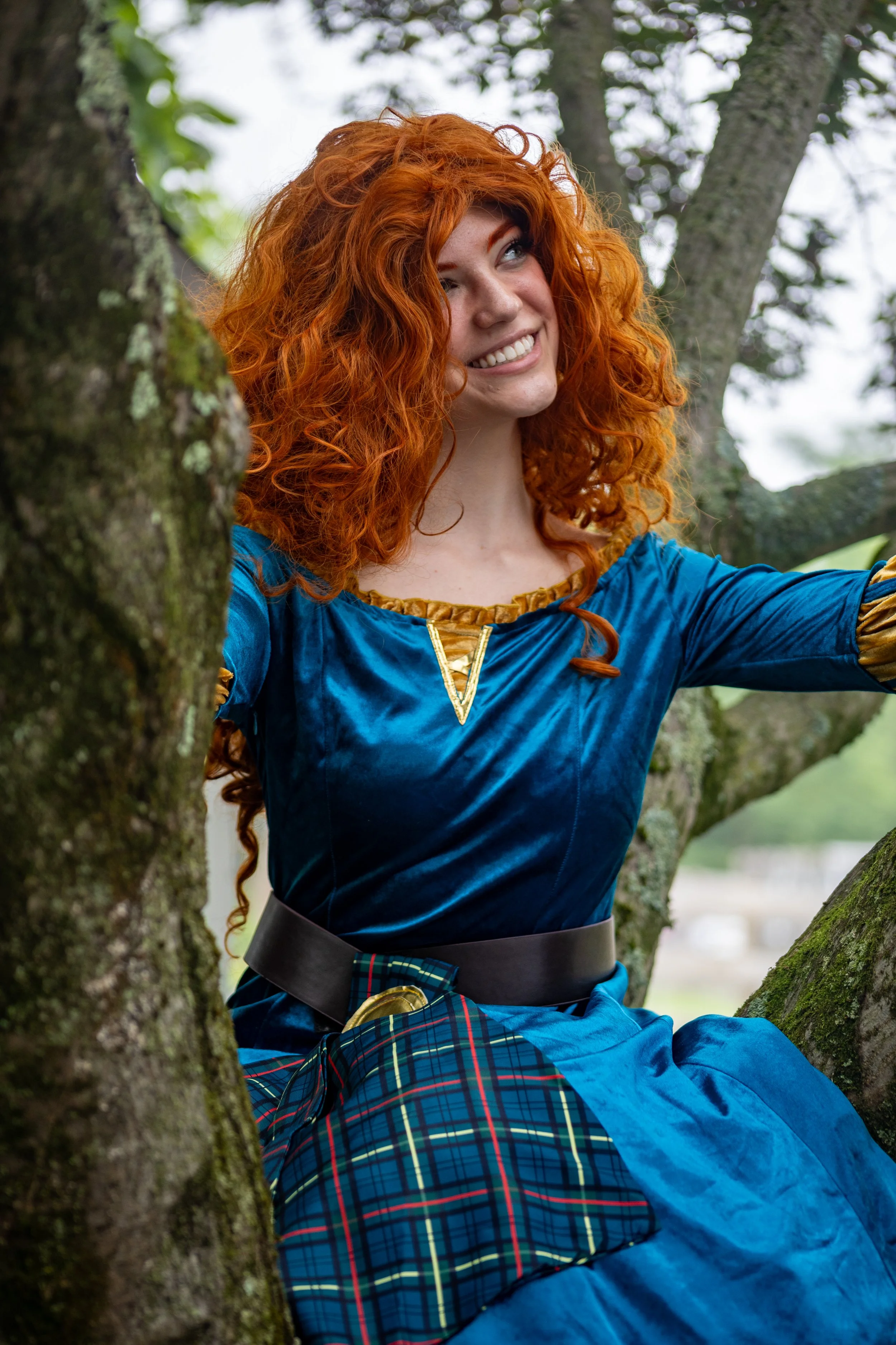 A woman with curly red hair wearing a blue dress and plaid skirt, sitting in a tree, smiling, outdoors.