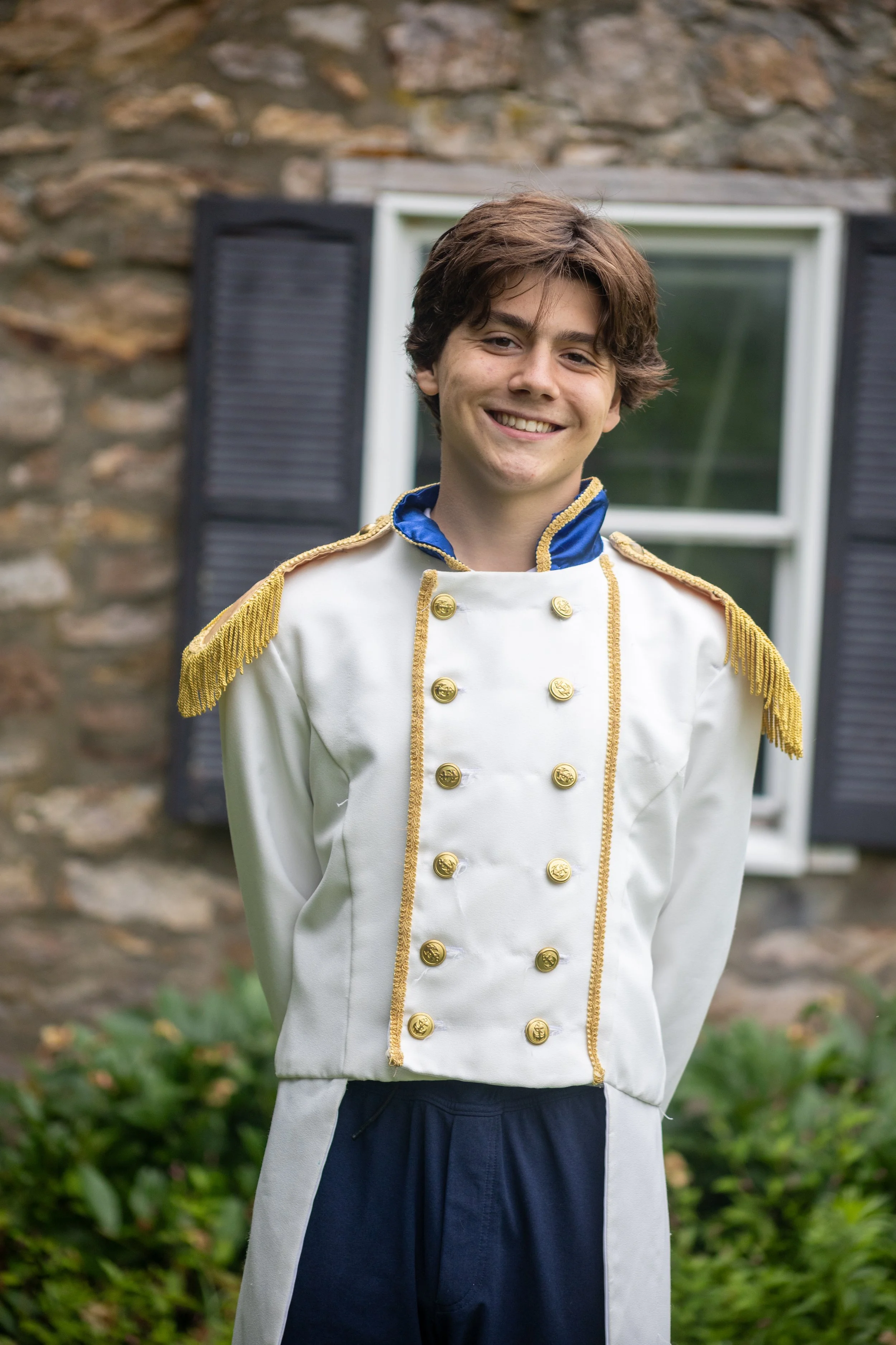 Young man dressed as Prince Charming with a white coat featuring gold trim and buttons, standing outdoors near a stone wall with a window and greenery.