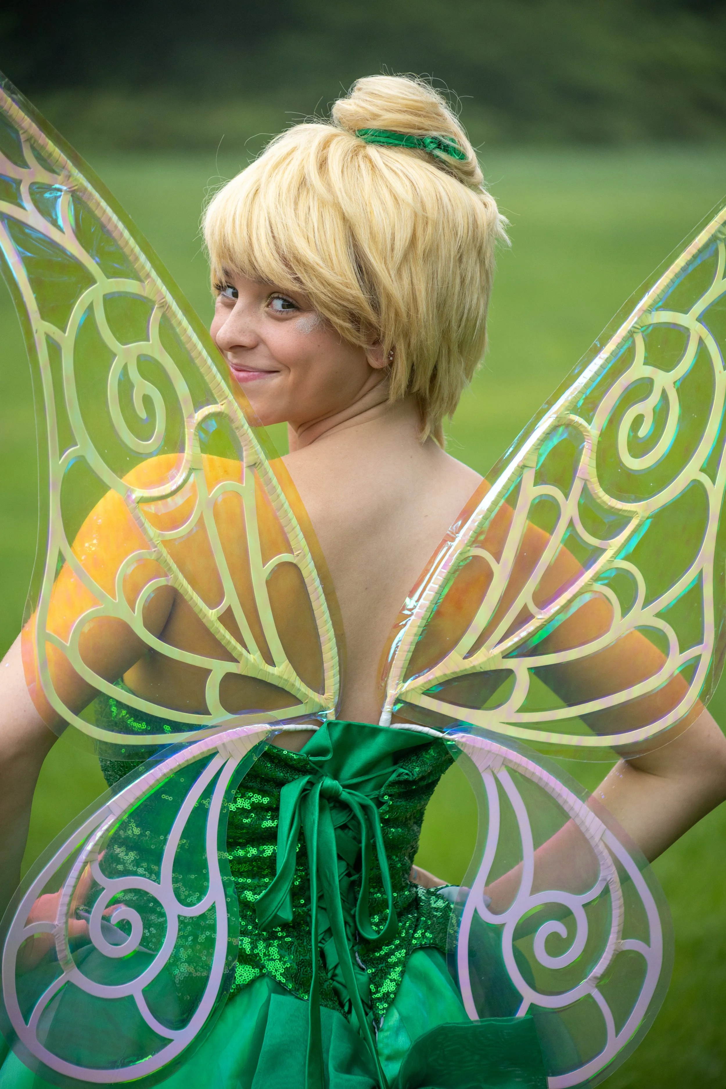A person dressed as Tinkerbell with large iridescent butterfly wings, blond hair with a green hair tie, wearing a sparkly green dress, smiling outdoors on a grassy background.