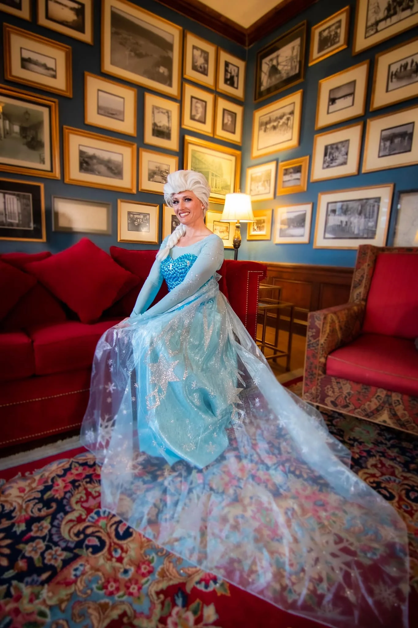 A woman dressed as Elsa from Disney's Frozen, sitting on a red sofa in a cozy room with blue walls decorated with many framed black and white photographs, and a lamp beside her.