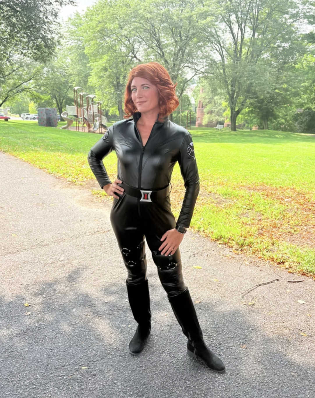 Woman with red hair standing outdoors in a park, wearing a black, shiny, tight-fitting leather jumpsuit and boots, with trees and a playground in the background, inspired by Black Widow.