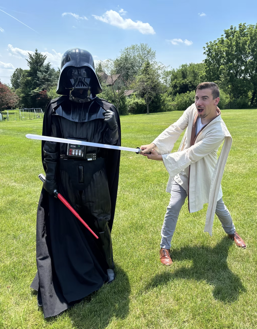 A man dressed as Obi-Wan Kenobi is fighting Darth Vader dressed in black armor with a red lightsaber, in a grassy outdoor area on a sunny day.