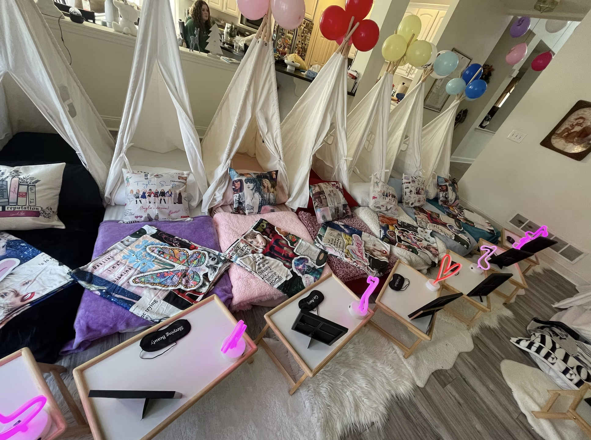 A room decorated for a girls' sleepover or birthday party with beds arranged in a row, each with a white canopy and colorful pillows featuring images of celebrities and pop culture. Small tables holding neon pink heart and musical note lights and eye
