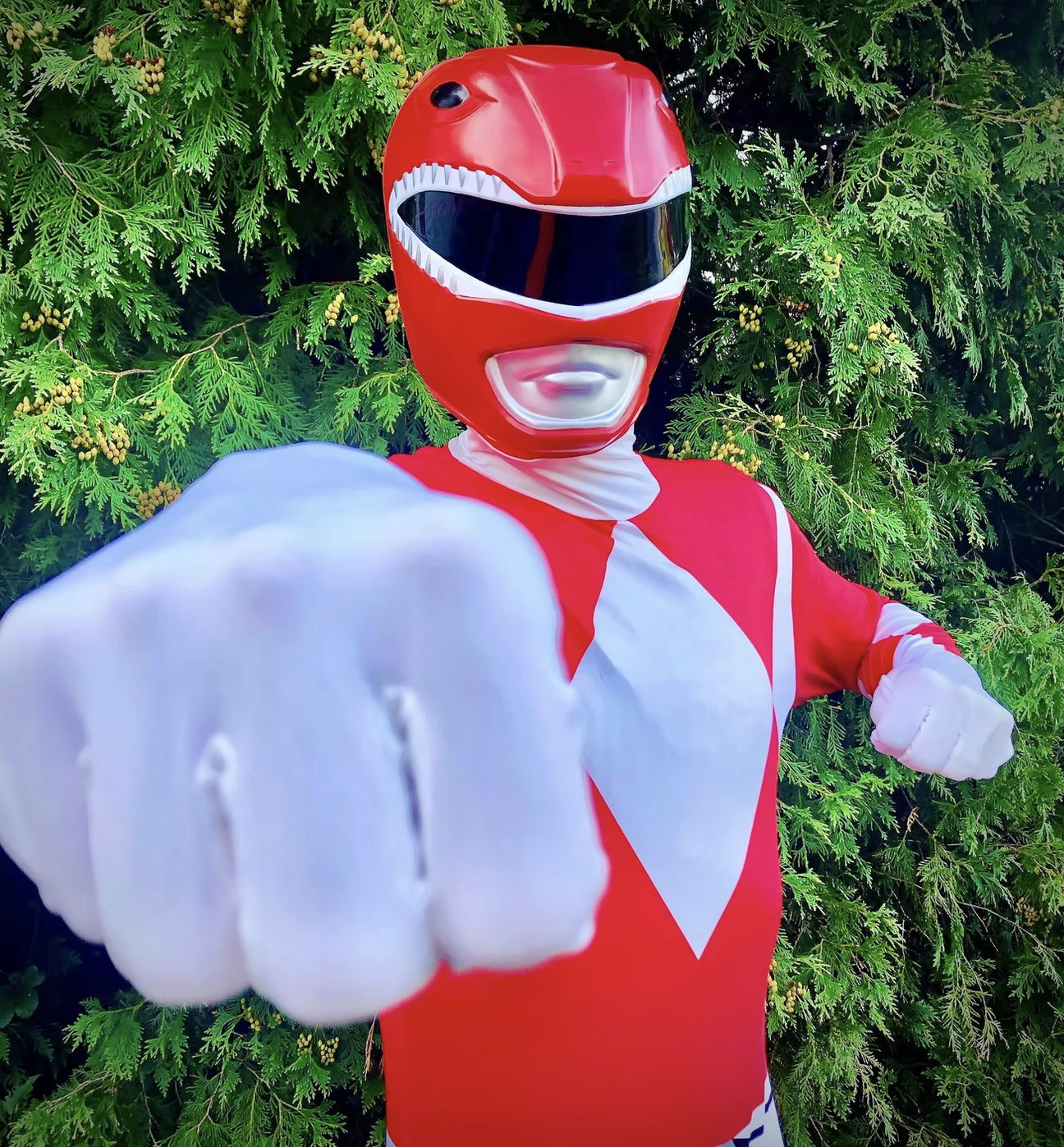 Person dressed as a red Power Ranger superhero, pointing towards the camera, standing in front of green foliage.