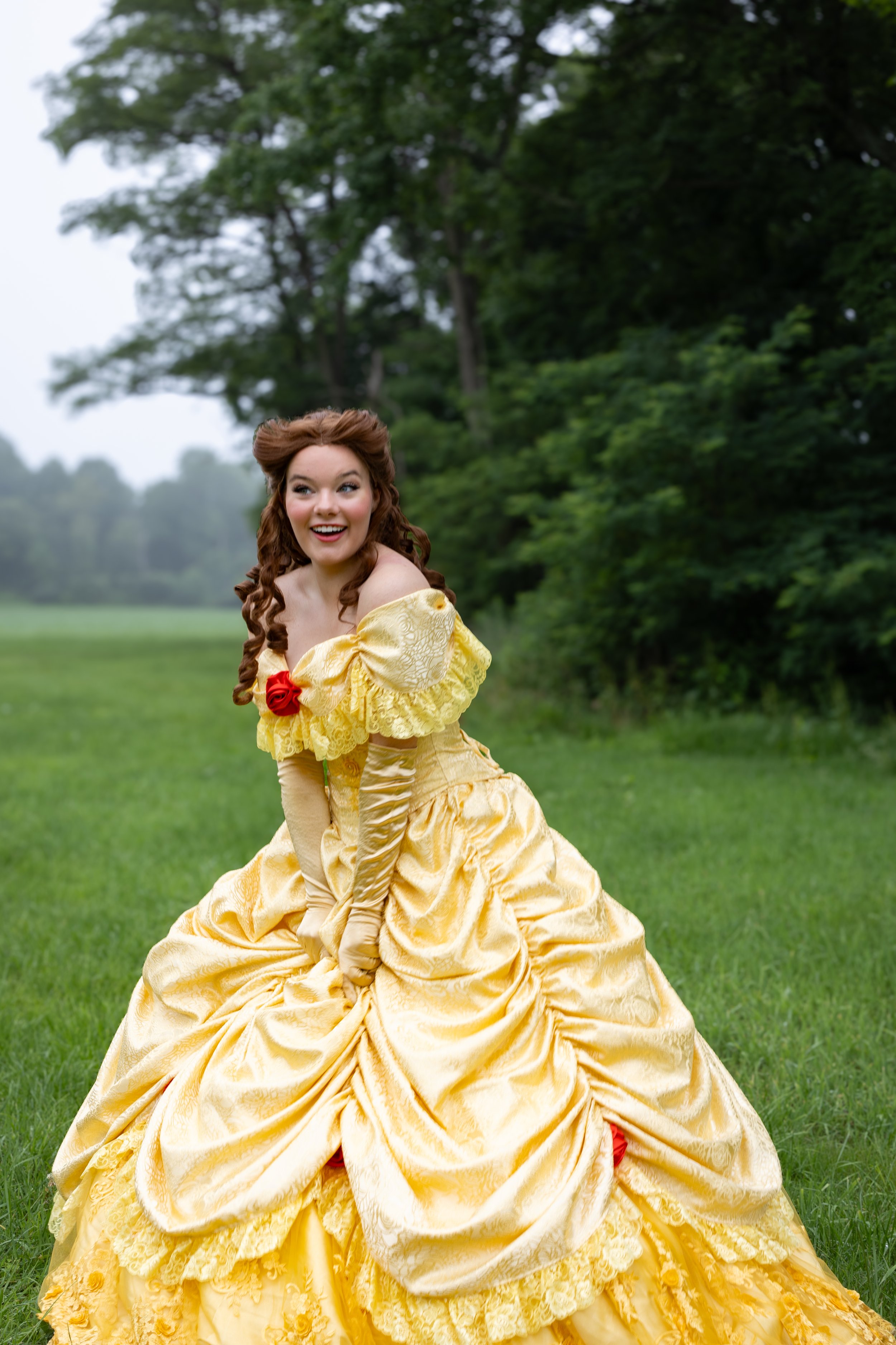 A woman dressed as Belle from Beauty and the Beast in a yellow gown, posing outdoors on a grassy field with trees in the background.
