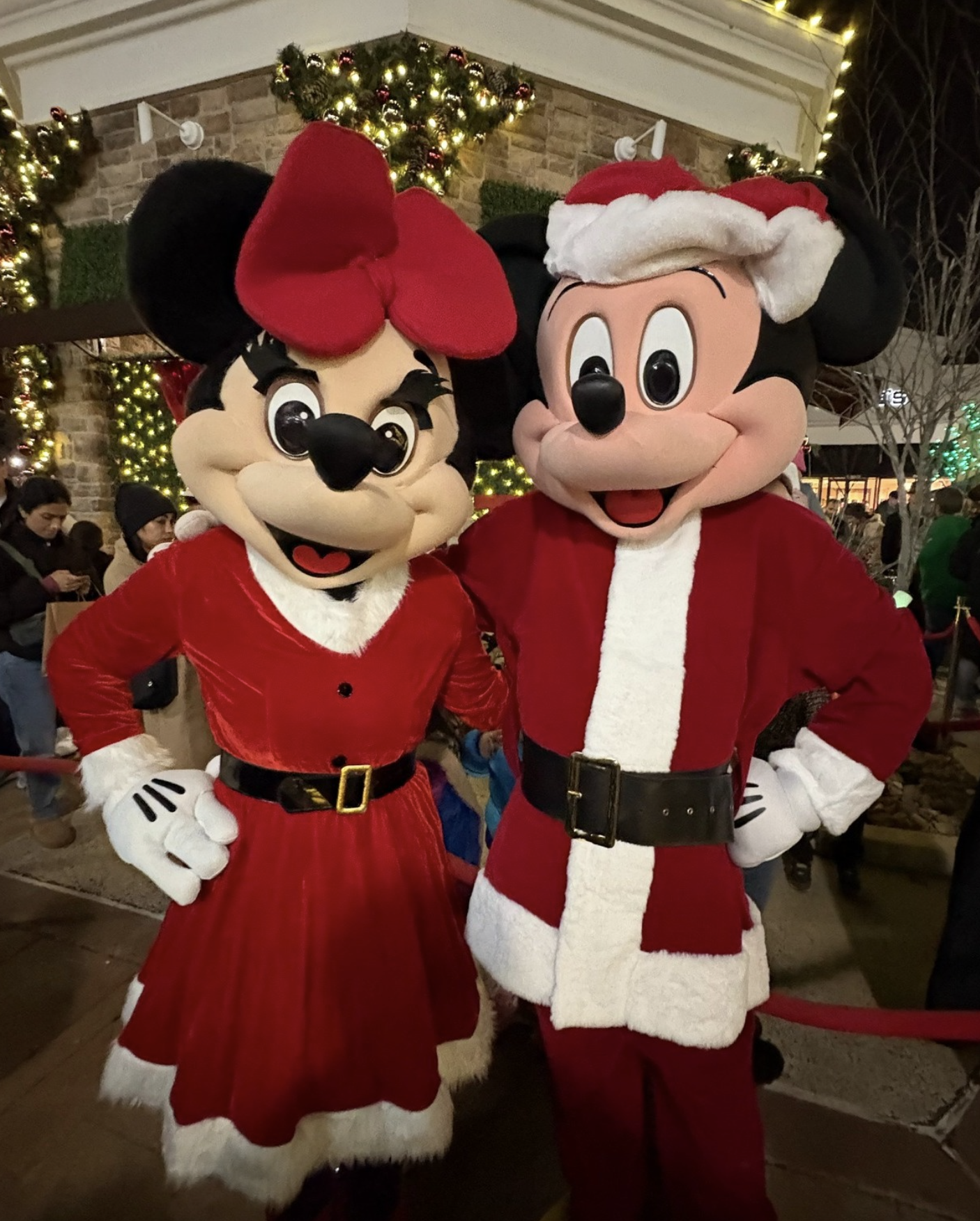 Minnie Mouse and Mickey Mouse dressed in Christmas costumes, standing together at a holiday event with decorated Christmas lights and people in the background.