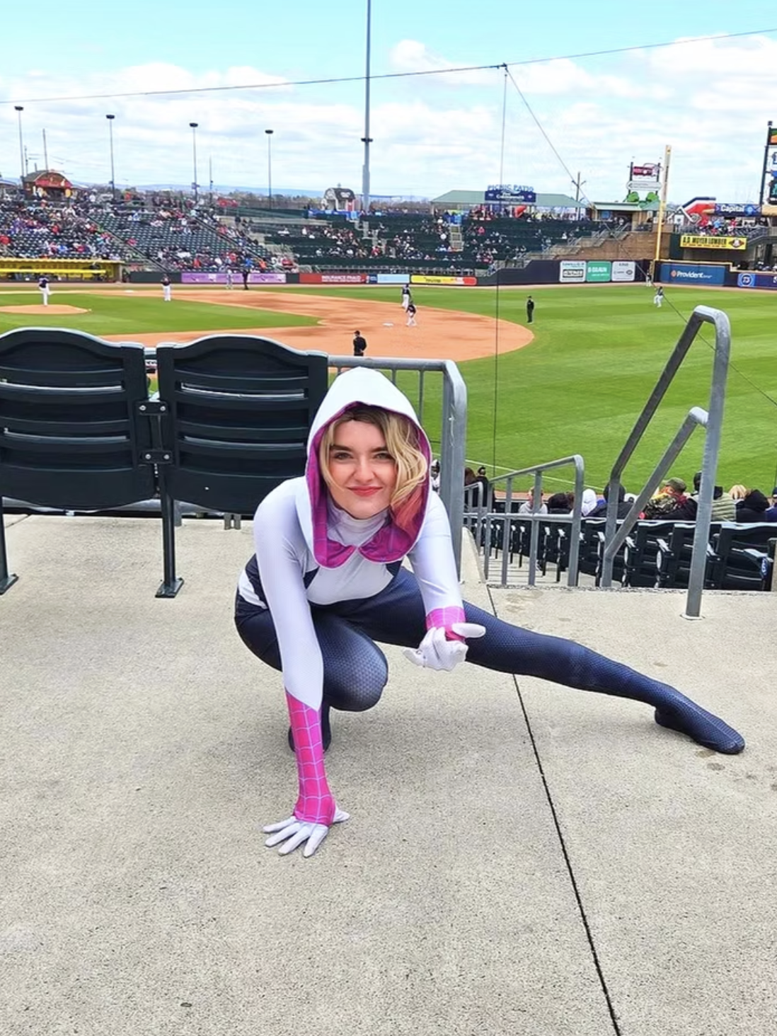 A woman dressed as Spider girl, posed on the ground with a baseball field in the background.