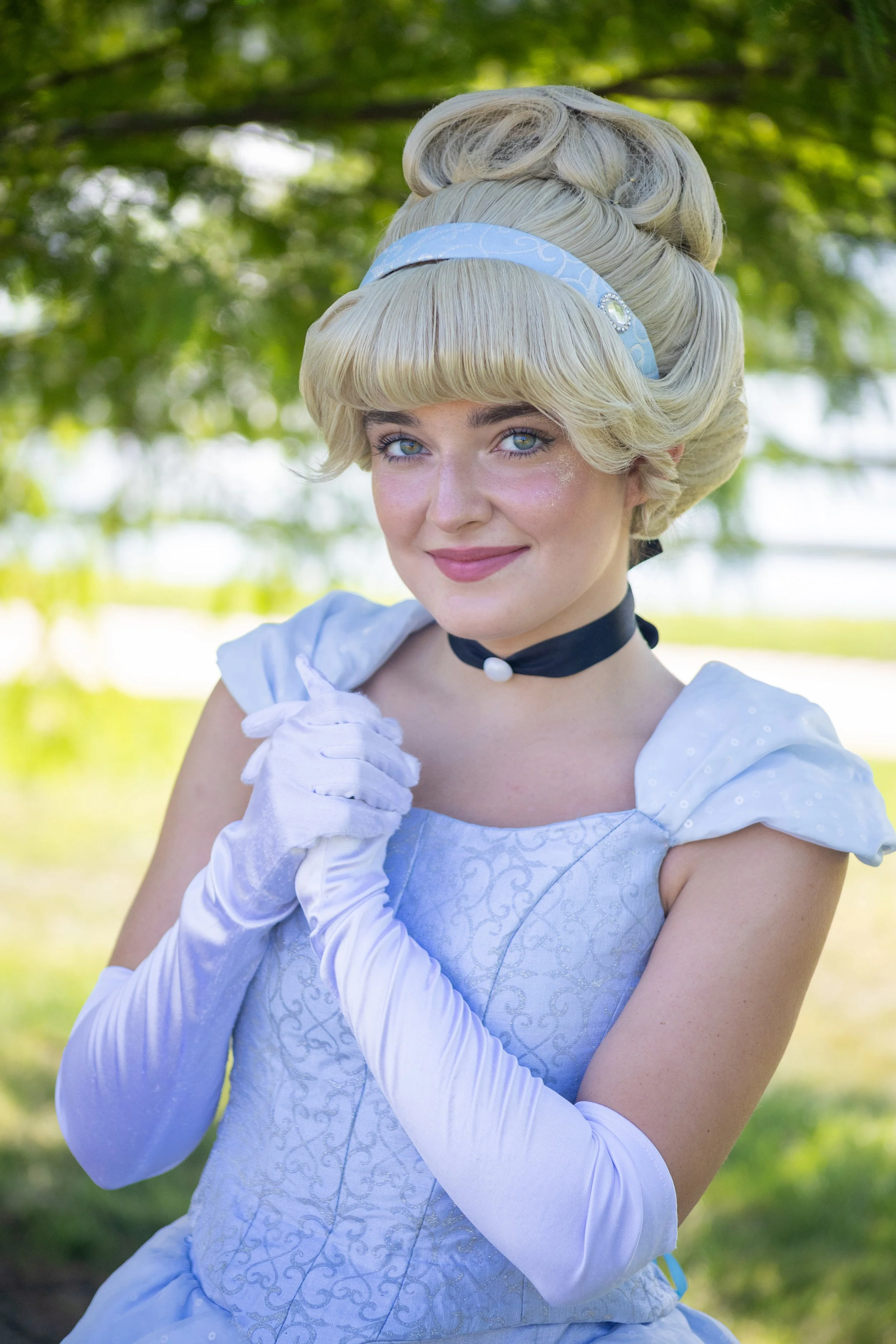 A woman dressed as Cinderella with a light blue gown, long white gloves, and a blue headband, outdoors with green trees in the background.