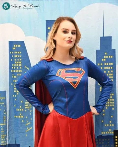 A woman dressed as Supergirl standing in front of a cityscape backdrop with buildings and clouds.