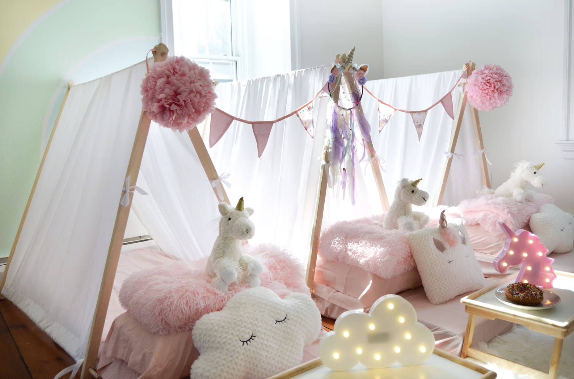 Cute unicorn-themed kids' bedroom decorated with plush unicorn toys, fluffy pink pillows, a glowing cloud-shaped light, and a pink-lit unicorn lamp, featuring a white canopy with pink accents and pastel-colored bunting for a whimsical, fairy-tale amb