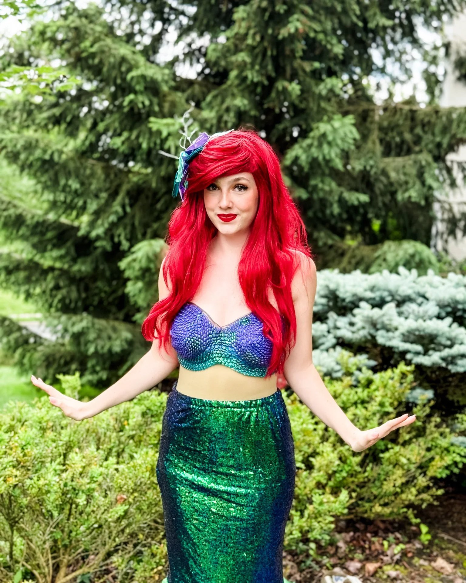 A woman dressed as a mermaid with long red hair, wearing a colorful, sequined mermaid top and a matching green-and-blue sequined skirt, standing outdoors in front of green trees and bushes.