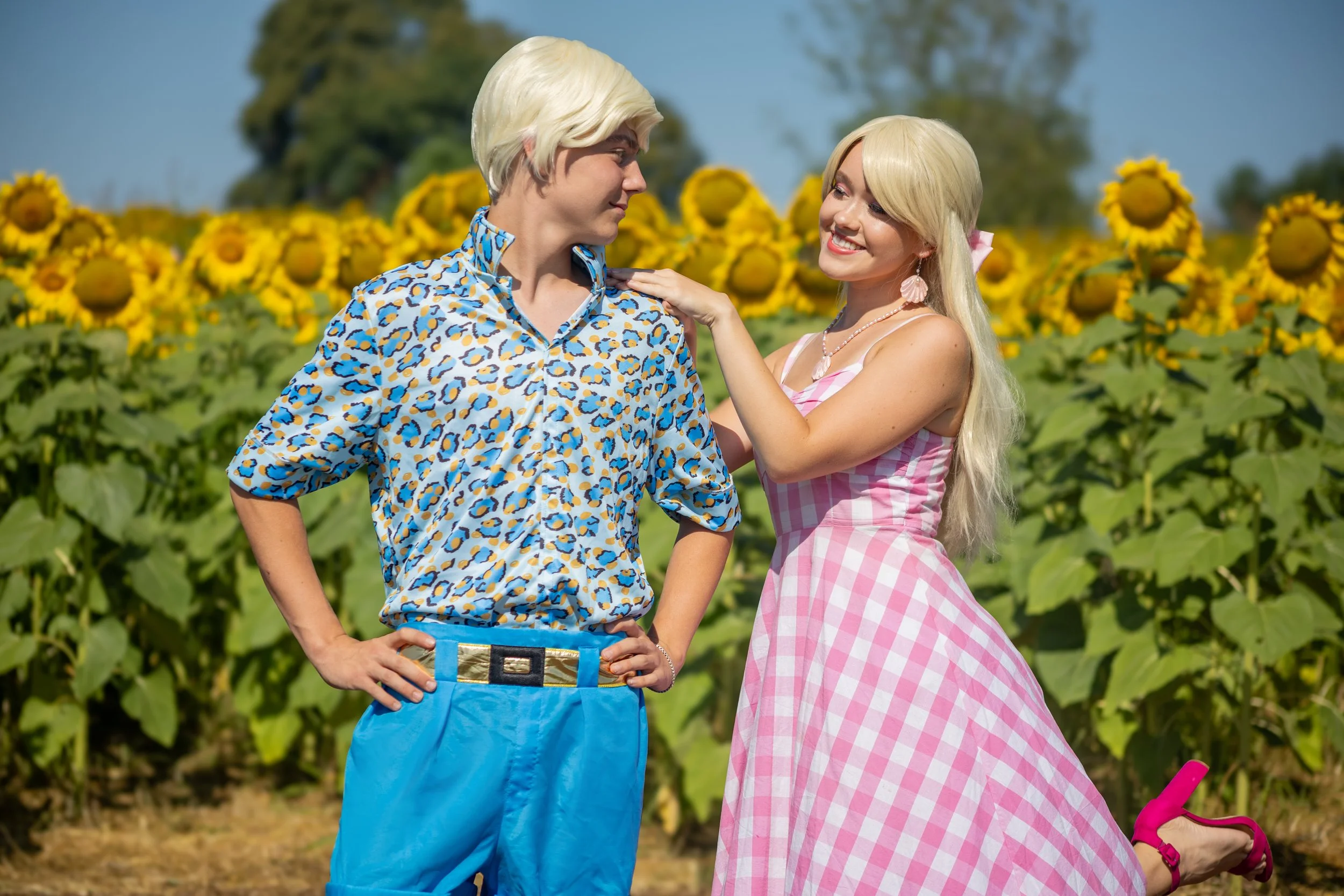 A man and woman in colorful clothing standing in a sunflower field, with the woman smiling and touching the man's shoulder, inspired by Barbie and Ken.