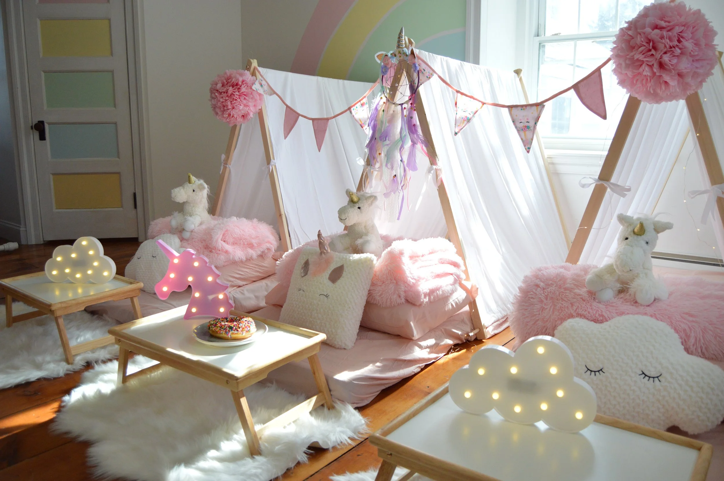 Pink and white unicorn-themed children's party setup with plush unicorn toys, cloud-shaped lights, bunting banners, pillows, and blankets inside a tent with sheer white curtains.