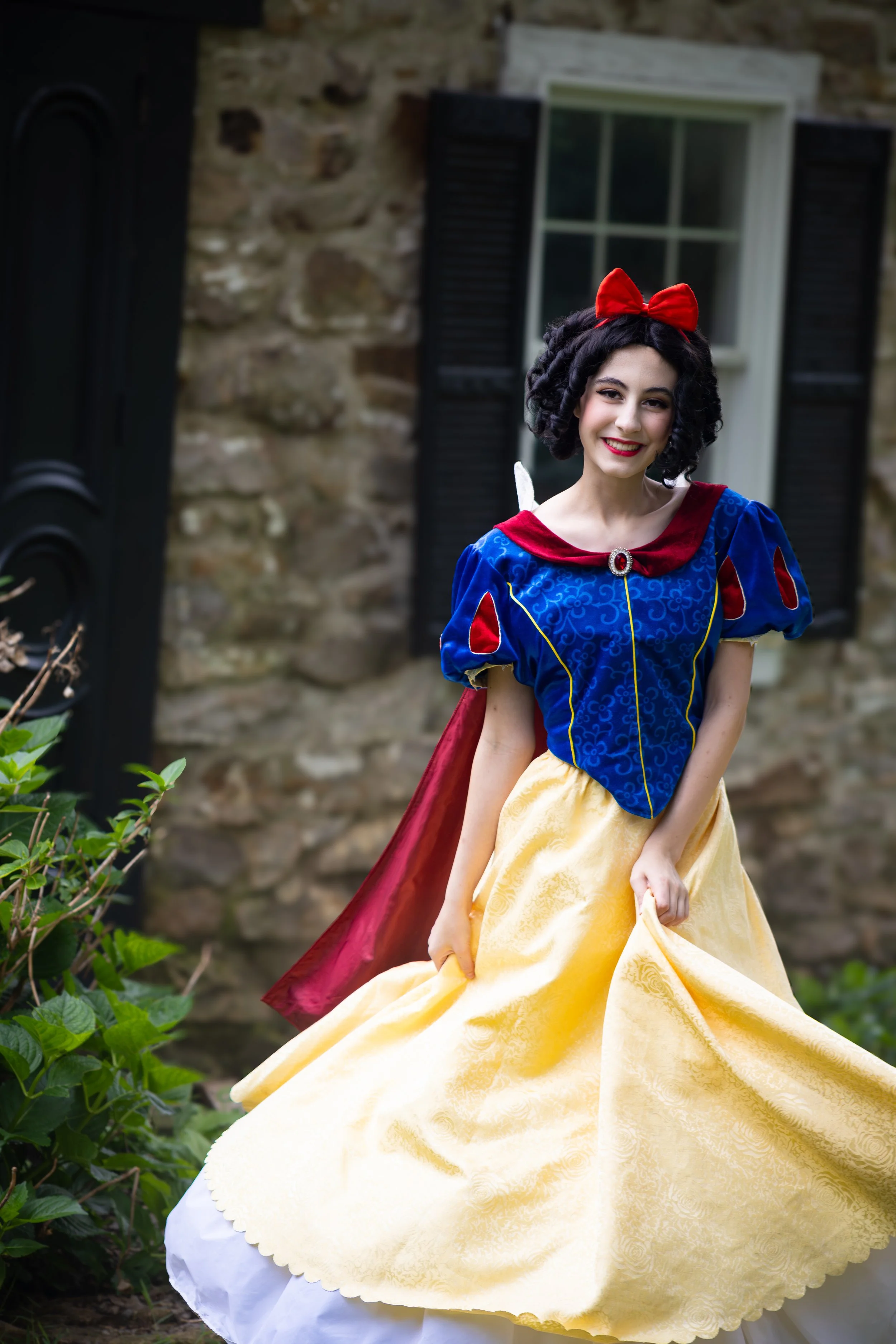 A young woman dressed as Snow White, wearing a blue and yellow gown with a red cape, a red bow in her black curly hair, standing outdoors in front of a stone building with a window, smiling at the camera.