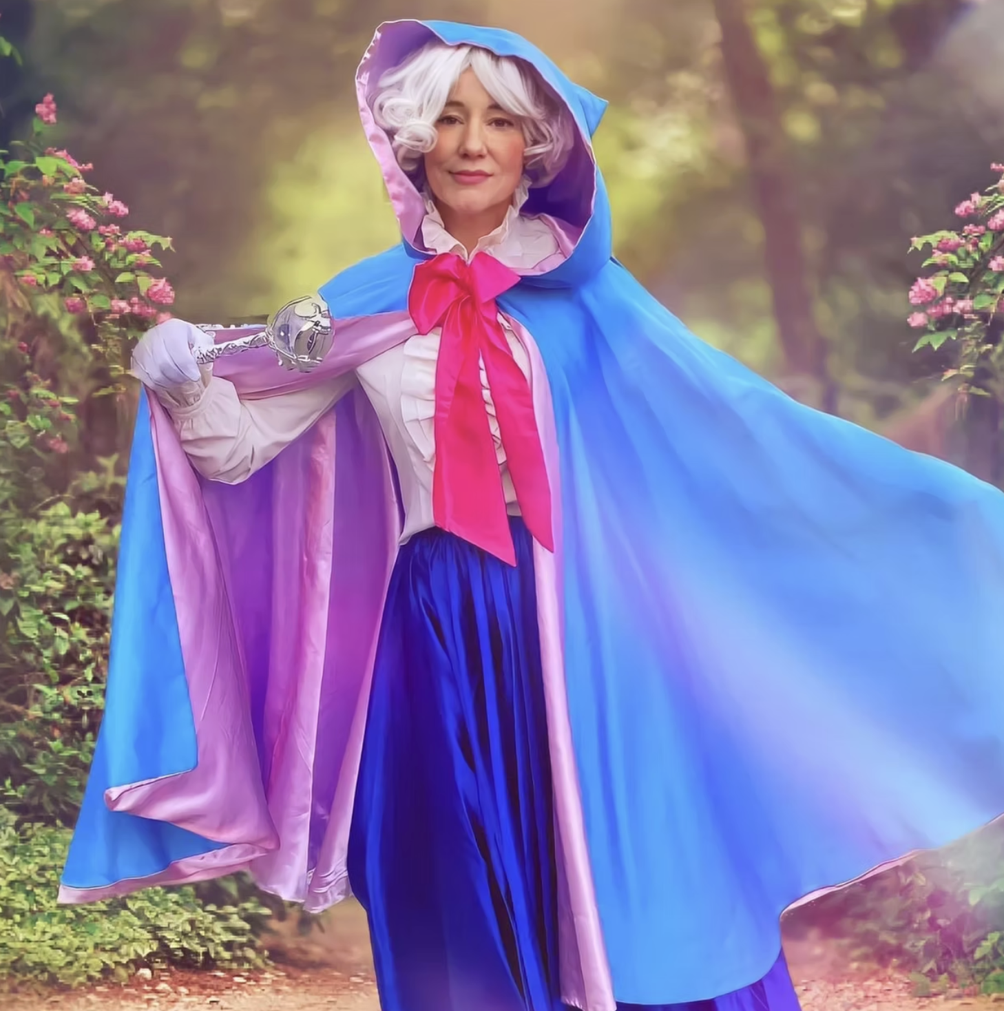 A woman dressed as the Fairy Godmother with a wand standing in a forest with pink flowers and green foliage.