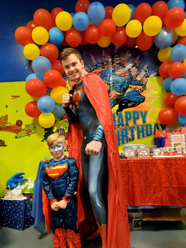 A young boy and a man dressed as superheroes at a birthday party. The boy is dressed as Superman with face paint, and the man is dressed as Superman or superman-like character. They are standing in front of a colorful balloon arch and a birthday table.