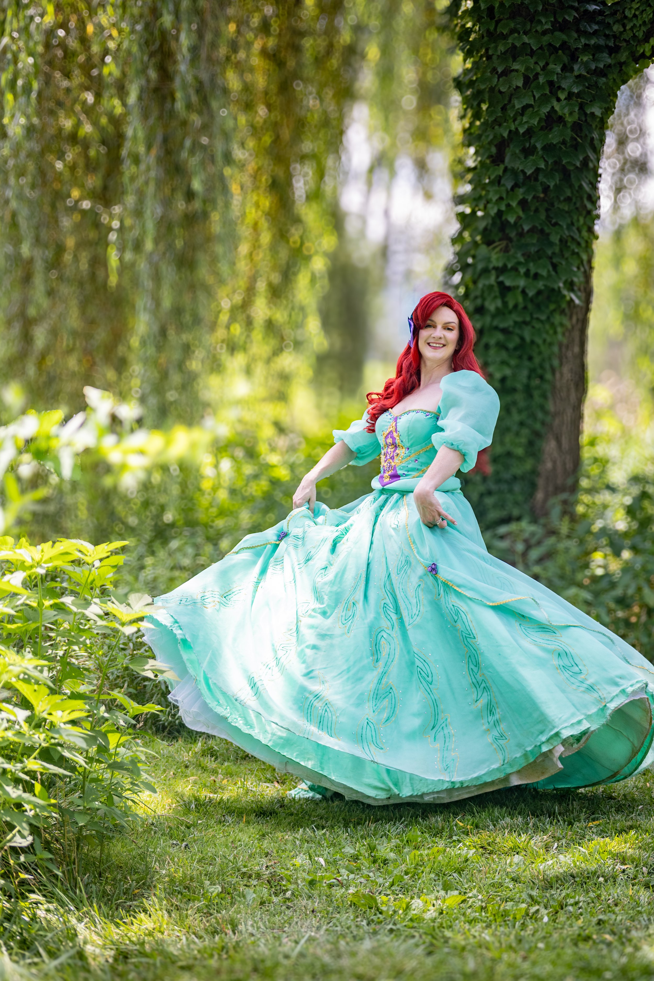 A woman with long red hair wearing a light blue, princess-style gown standing outdoors in a wooded area, smiling at the camera.