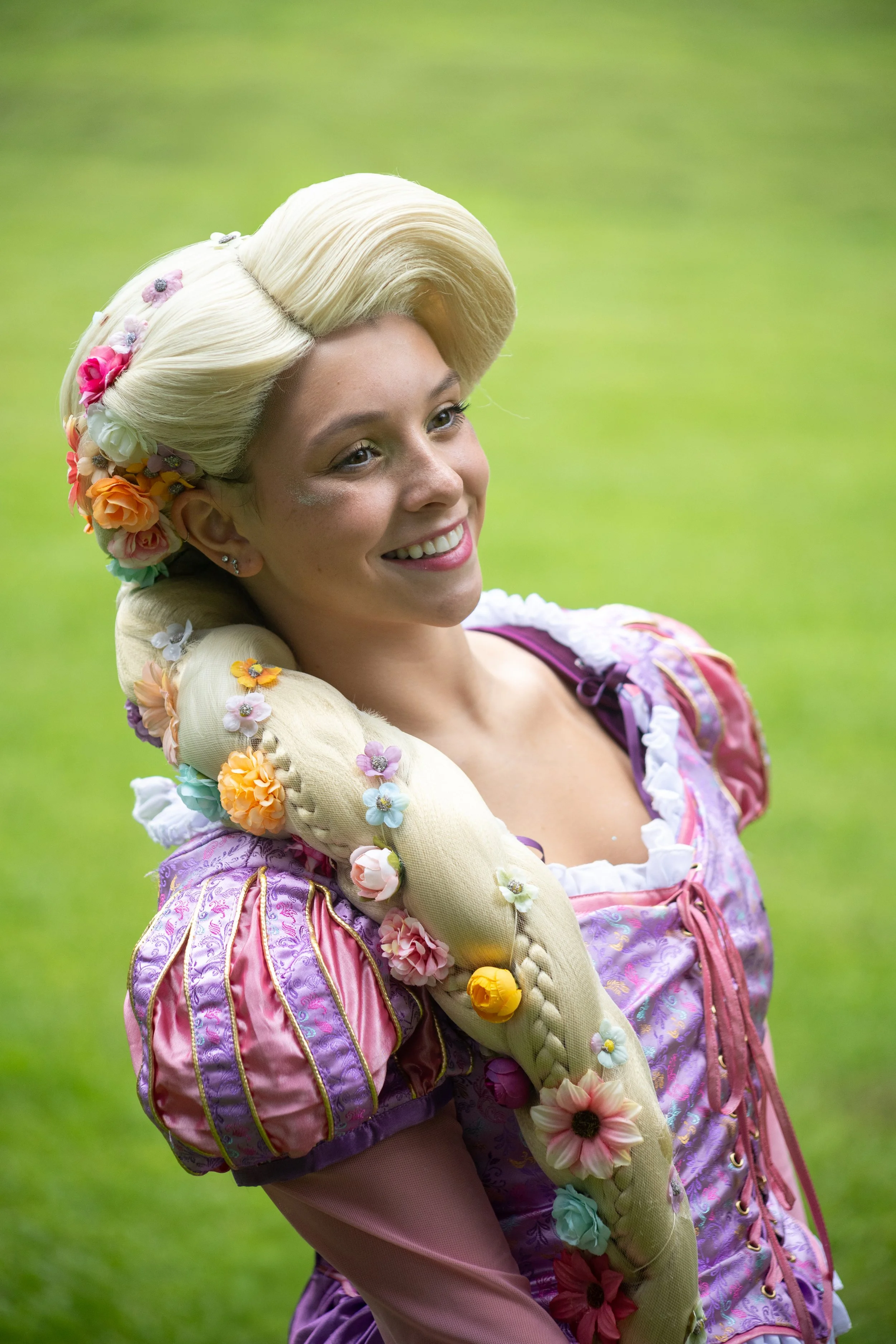 A woman dressed in a colorful, princess-like costume with a long braided hair decorated with flowers, standing on a grassy field and smiling