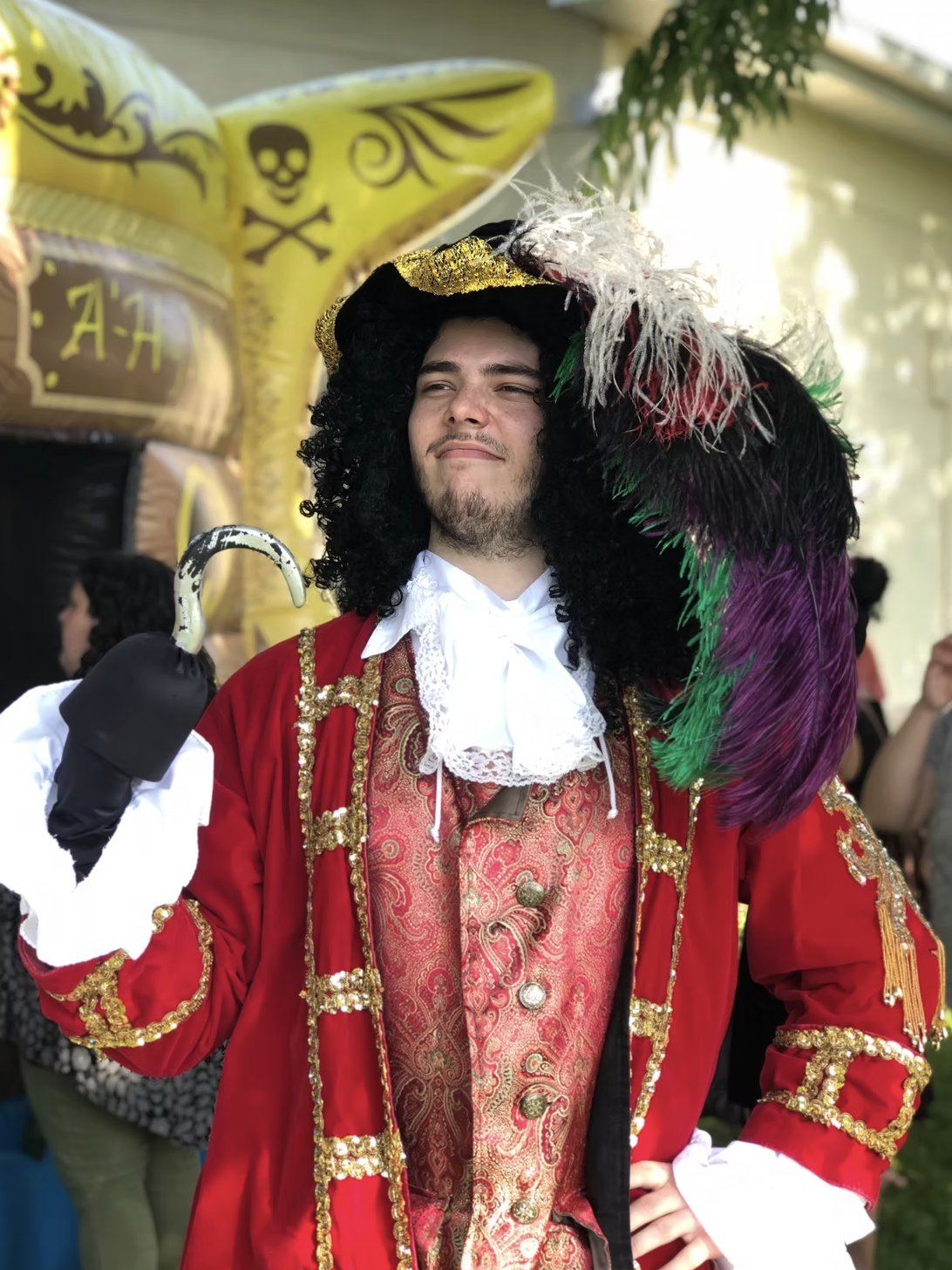 Person dressed as a pirate with a black curly wig, red jacket with gold embroidery, and a large feathered hat, standing outdoors during a festival or parade.