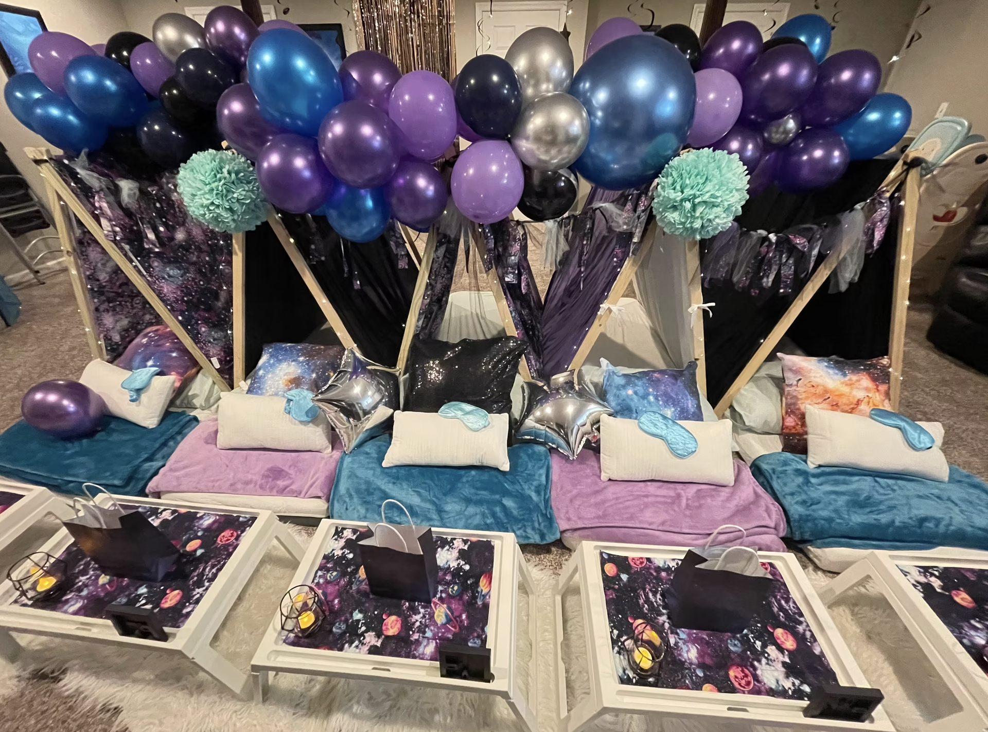 Cosmic-themed party setup with galaxy-patterned cushions, star-shaped balloons, and balloon arrangements featuring purple, blue, black, and silver balloons. Small tables with decorative space-themed mats and gift bags are in front.