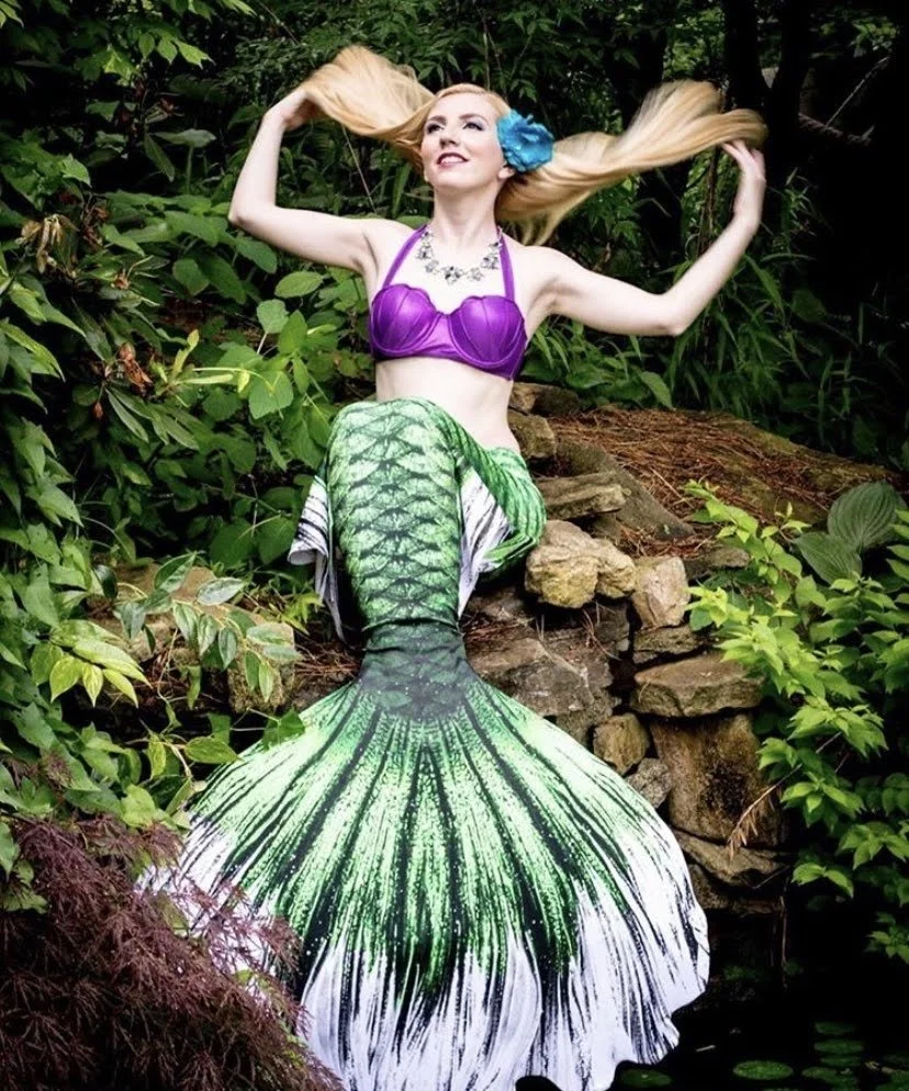 Woman with long blonde hair wearing a purple top and large blue flower hair accessory, sitting outdoors among green foliage and rocks, with a mermaid tail in green and white colors.
