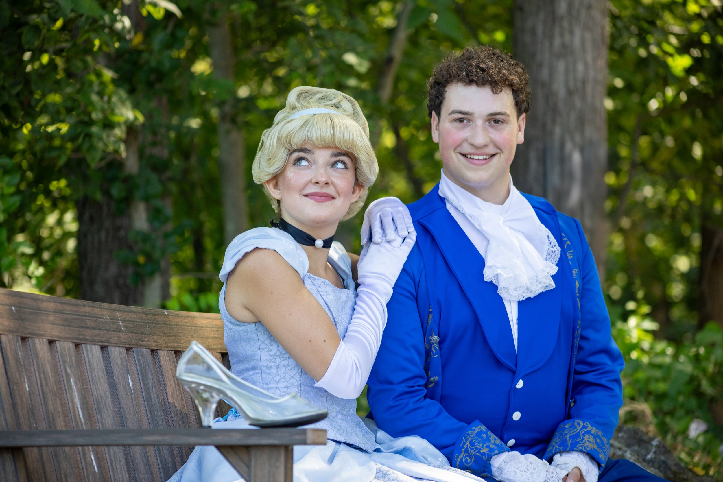 A woman and a man dressed in historical costumes sitting on a park bench with trees in the background. The woman, with blonde hair styled in a vintage look, is wearing a light blue dress and white gloves, and has her hand resting on the man's shoulde