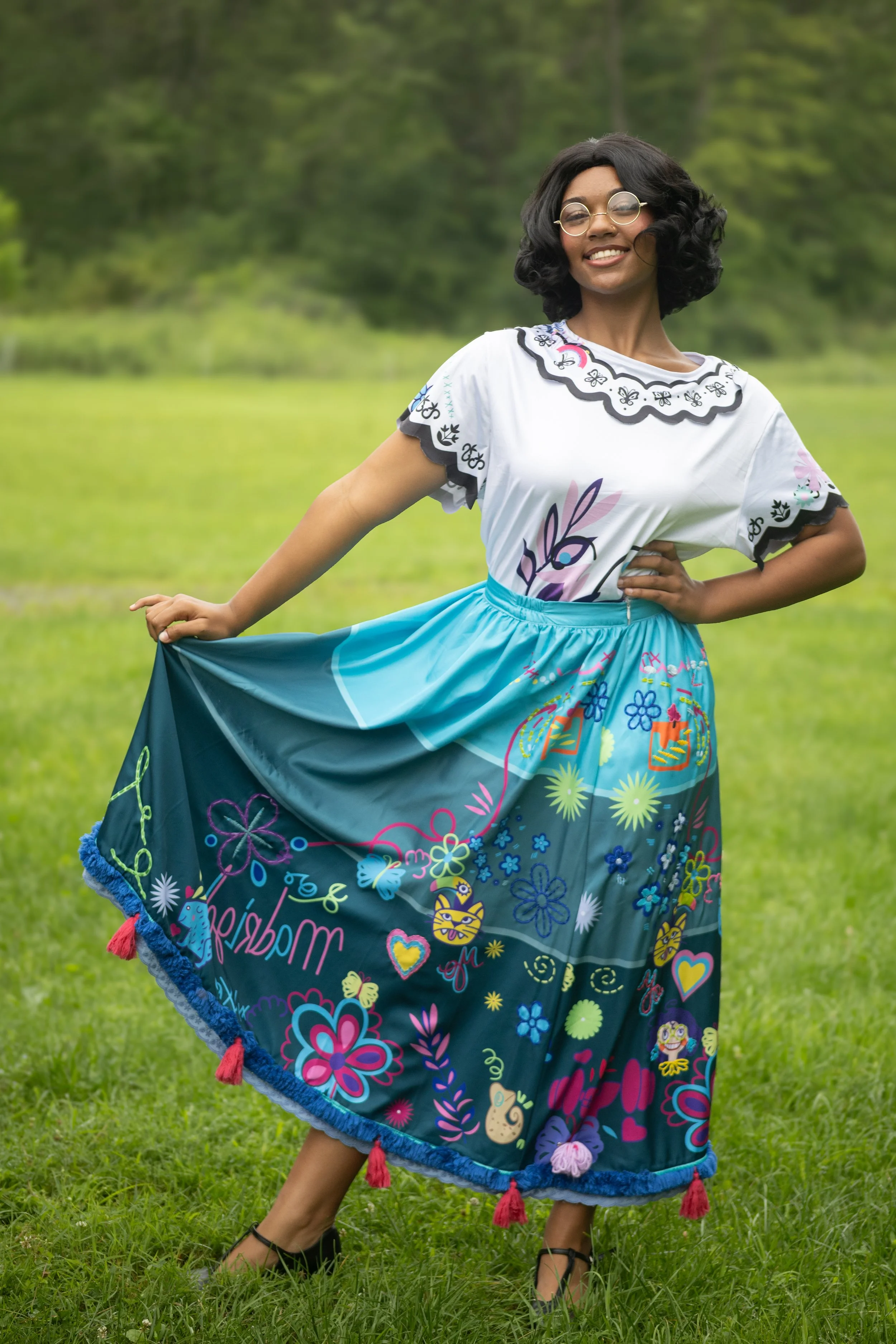 A woman standing in a grassy field, wearing a colorful skirt with floral and whimsical patterns, a white top with embroidered designs, glasses, and smiling, inspired by Encanto.