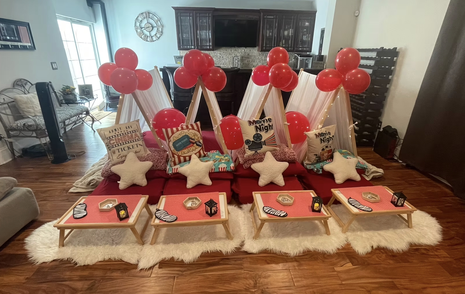 A cozy living room decorated for a movie night, featuring red balloons, star-shaped pillows, small tables with lanterns, and faux fur rugs.