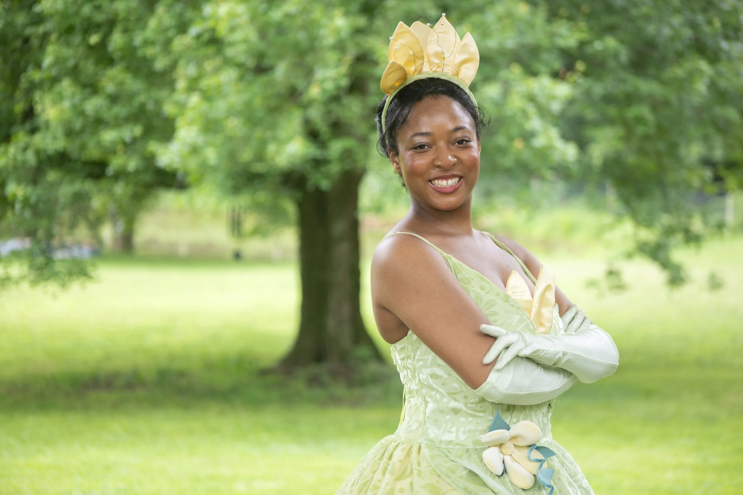 A woman in a yellow dress with a matching headband and gloves, smiling outdoors in a green park.