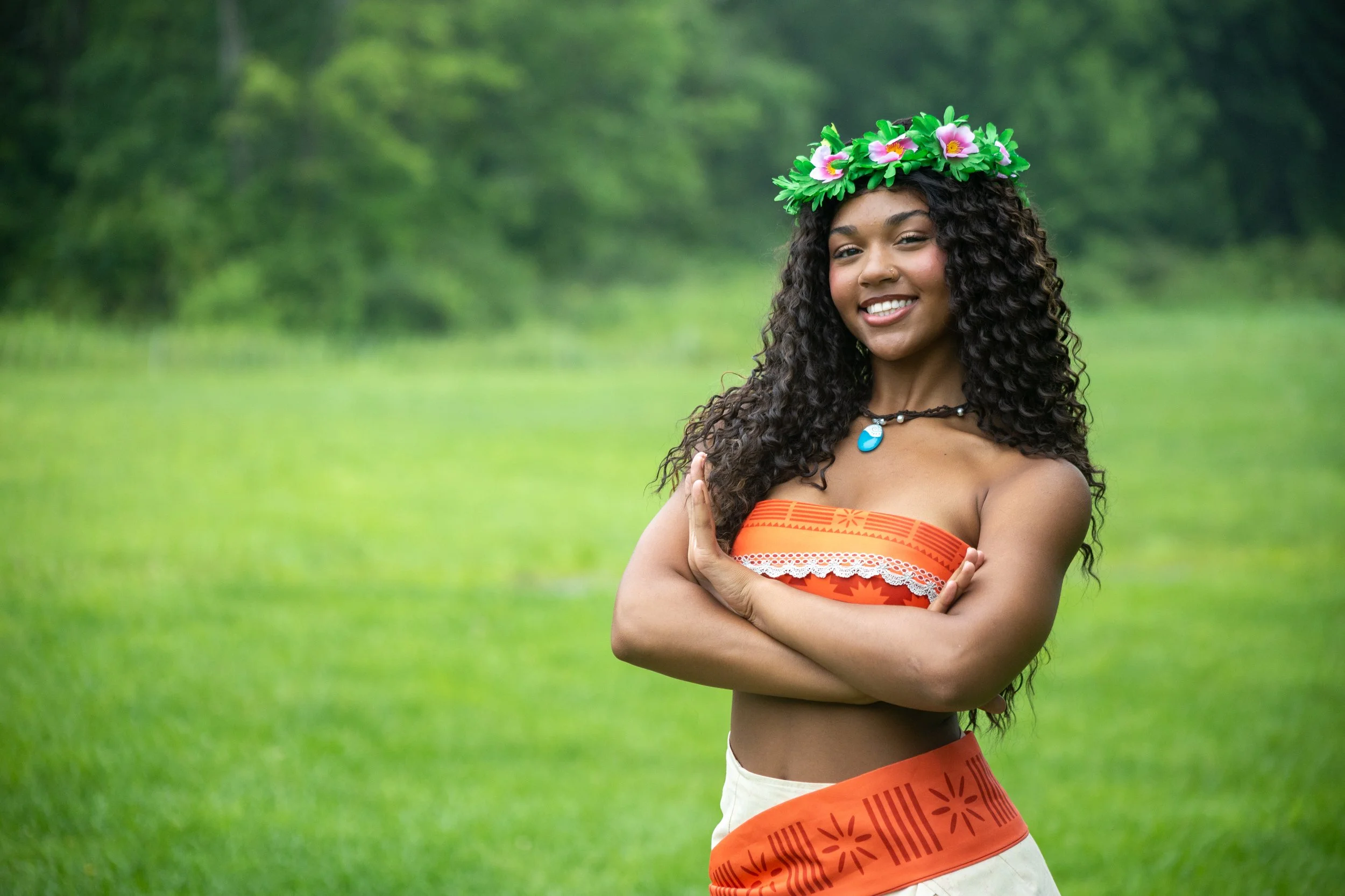 A young woman with long curly hair wearing a flower crown, an orange strapless top, and a necklace with a blue pendant, standing outdoors in a green grassy area.