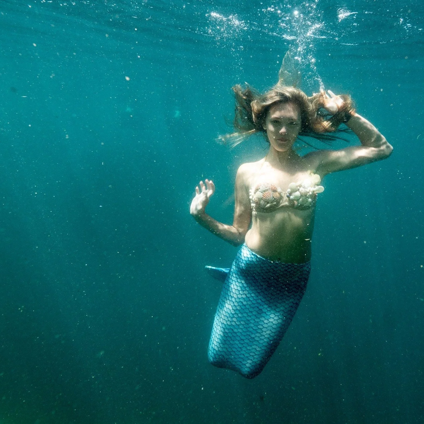 A woman dressed as a mermaid, with a shell bra and fish tail, is underwater.